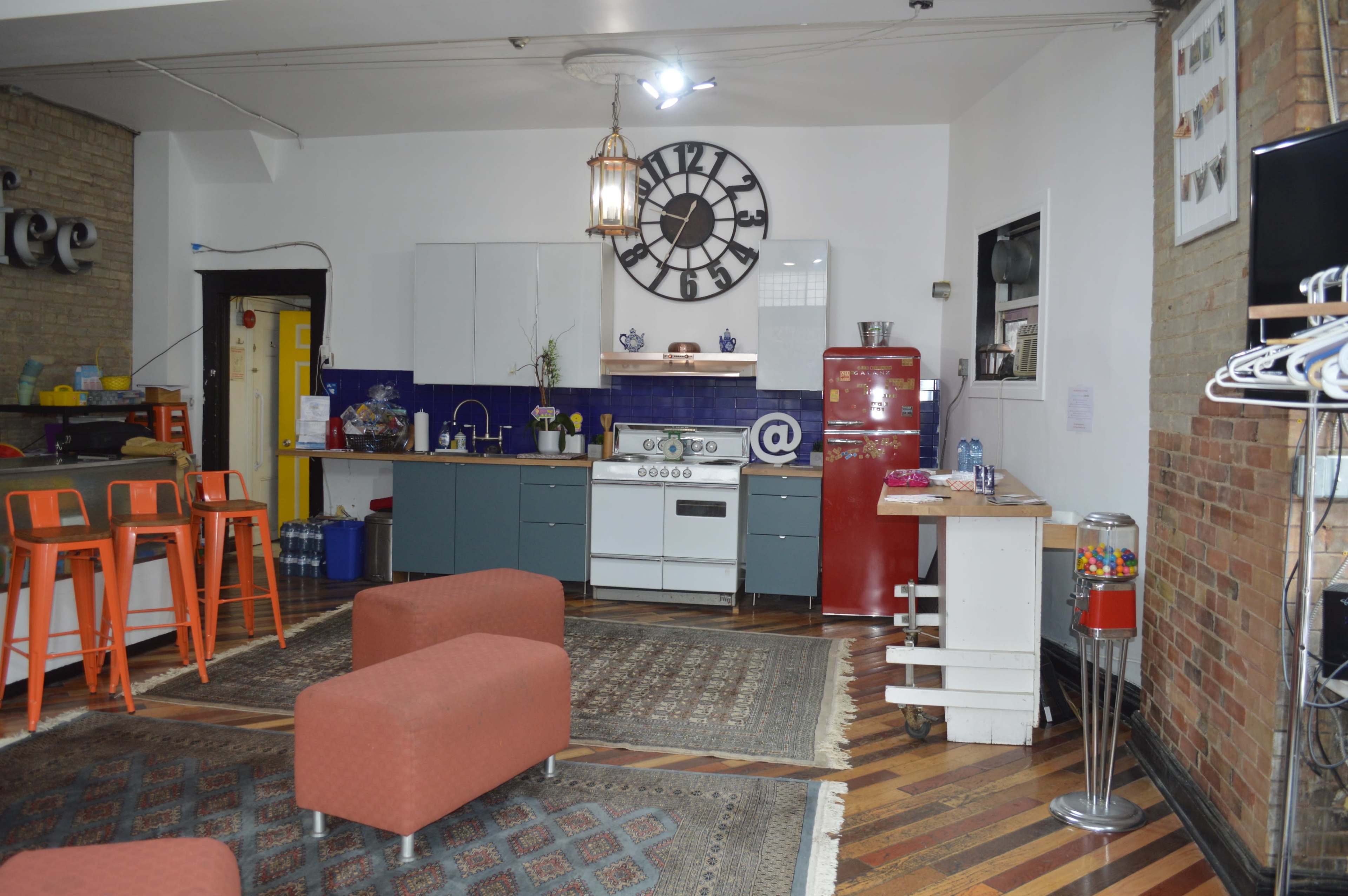 The image shows a kitchen area with a large wall clock, a red refrigerator, and orange bar stools, all set against a backdrop of exposed brick and wooden flooring.