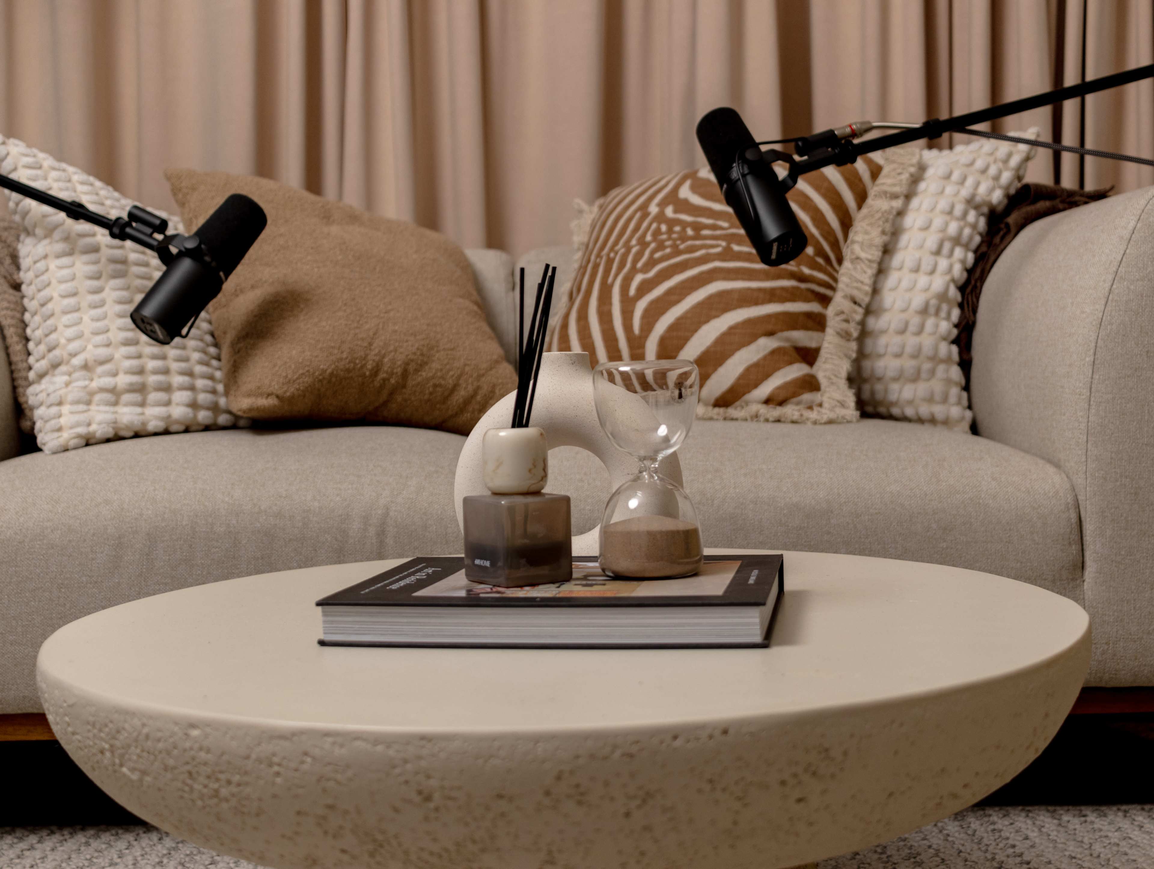 A modern living room features a light-colored sofa with various textured pillows and two microphones positioned above a coffee table displaying an hourglass, candles, and a stacked magazine.
