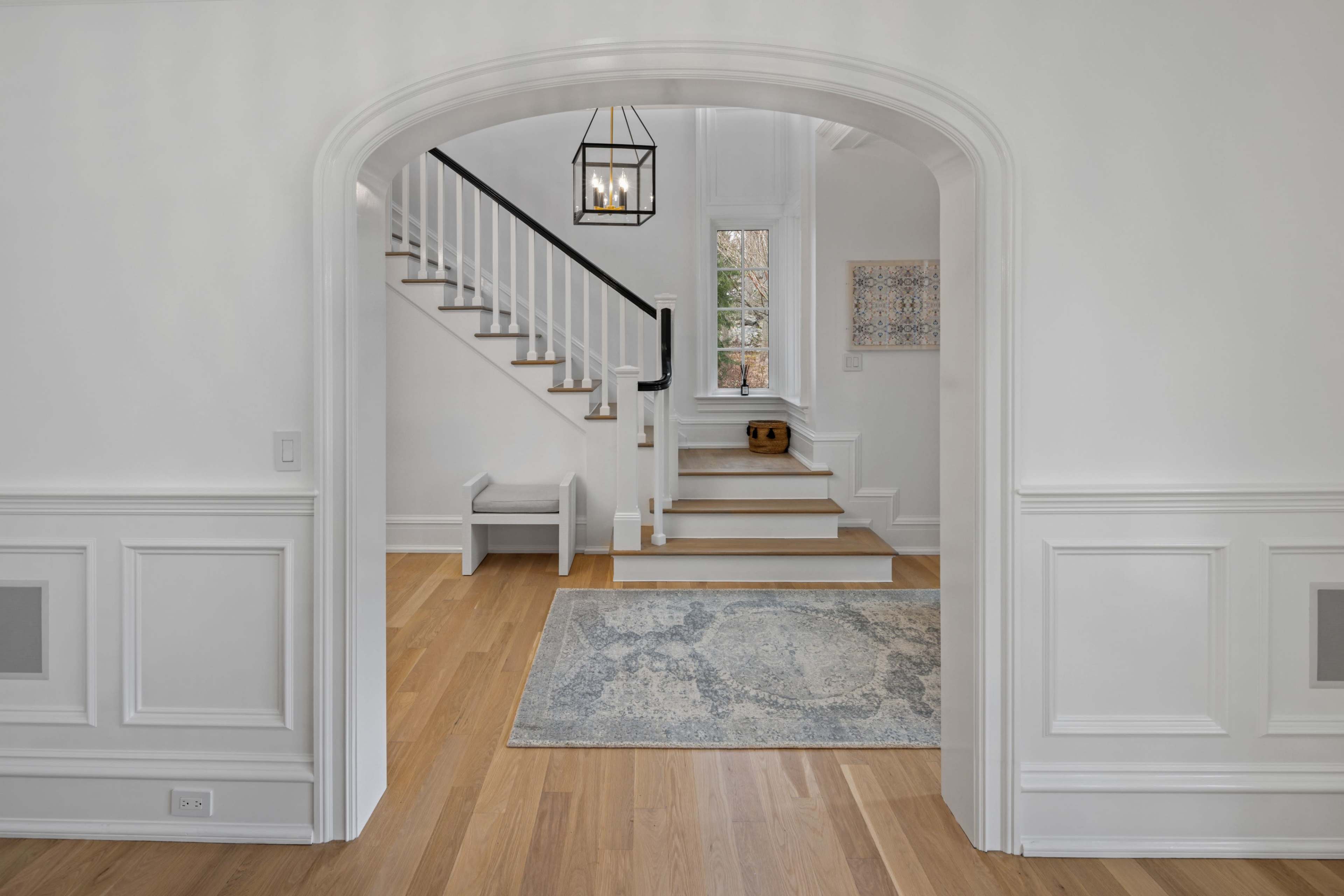 A spacious entryway features a staircase, a geometric light fixture, and a decorative rug on polished hardwood flooring.