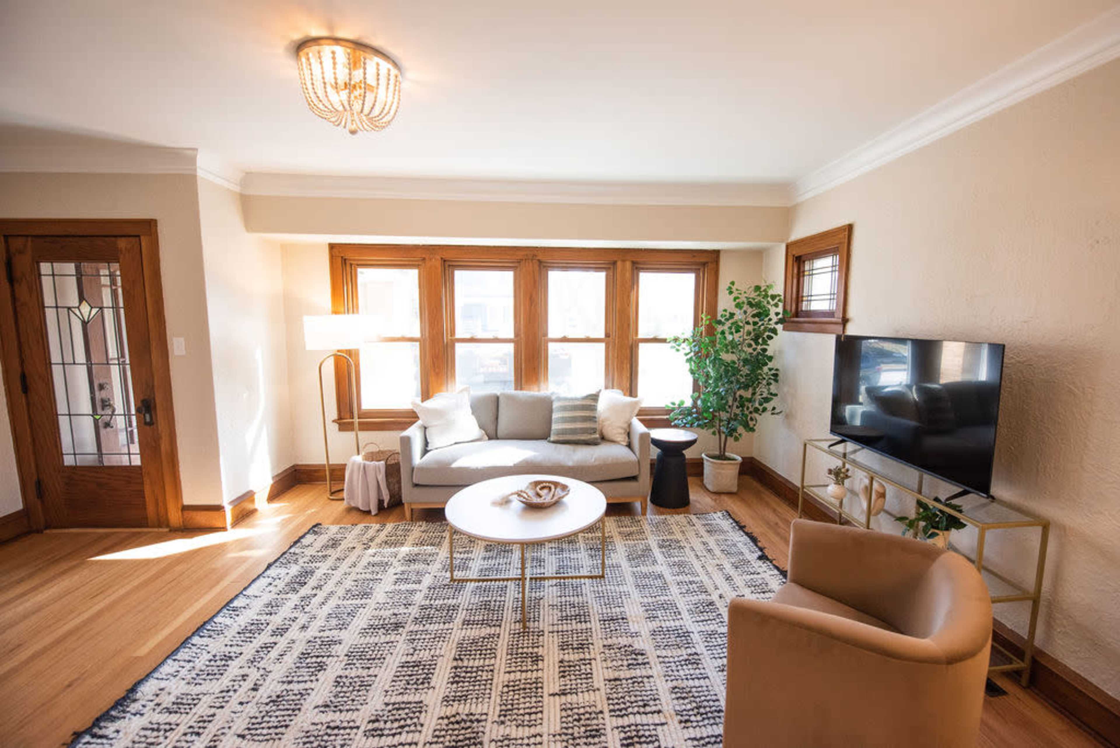 A well-lit living room features a couch, a coffee table, a chair, a television, and a small plant near a window.