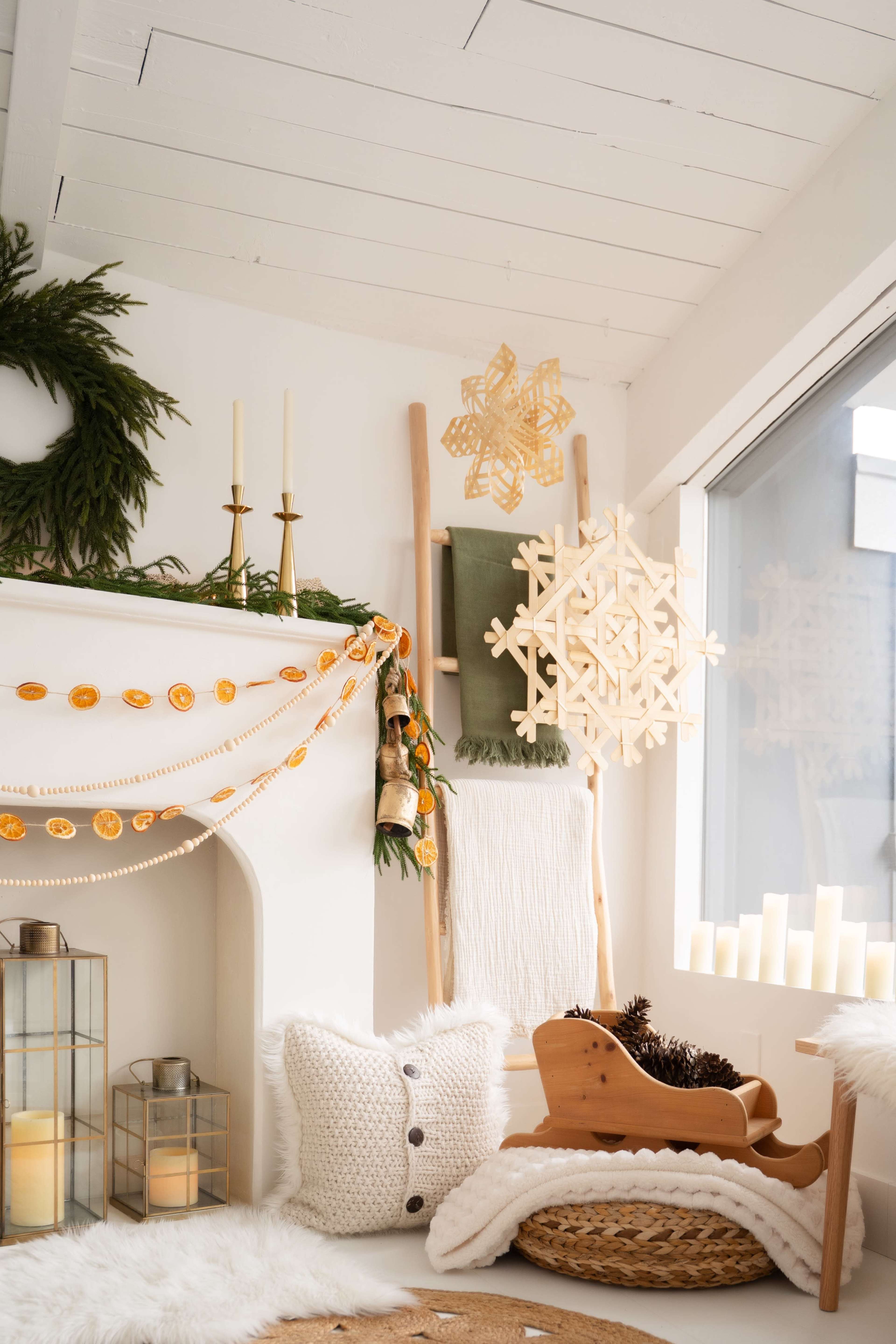 A cozy room features a white fireplace adorned with greenery, candles, and decorative garlands, complemented by a snowflake wall hanging and warm lighting.