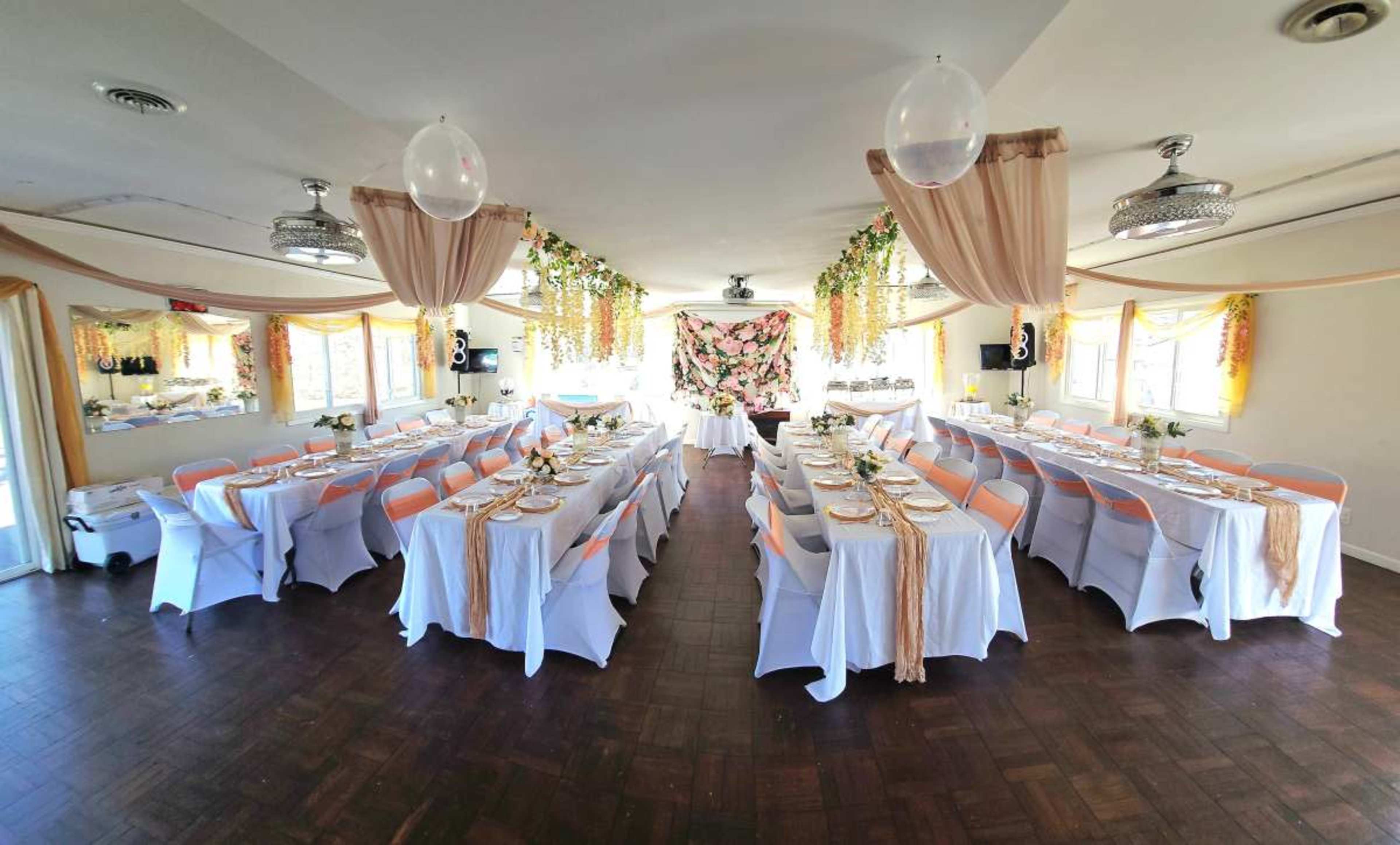 A banquet hall is set up with tables covered in white and orange cloths, adorned with floral centerpieces and draped fabric decorations overhead.