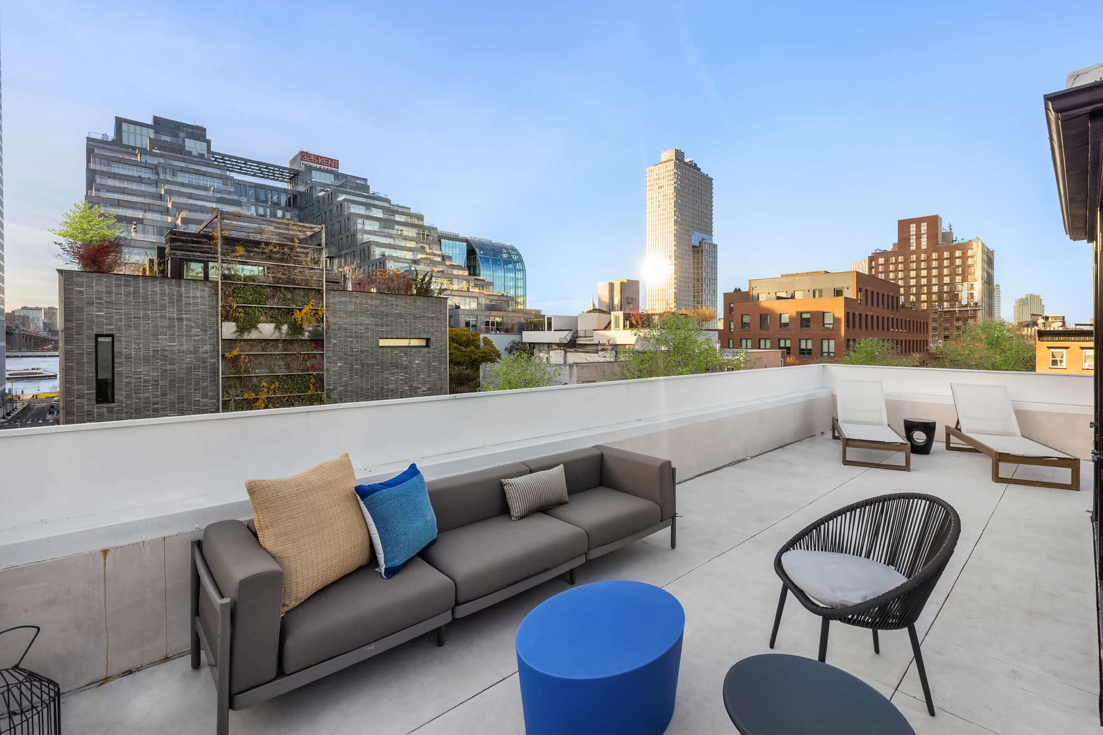 The image shows a rooftop terrace with modern outdoor furniture and a view of urban buildings and a clear sky.
