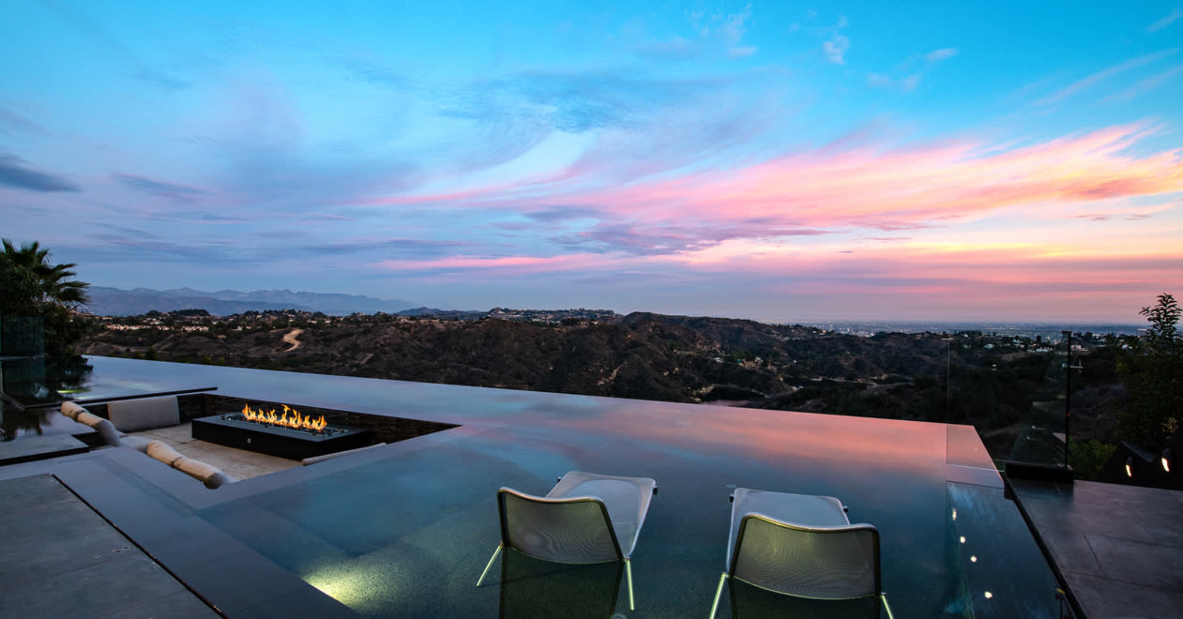 An infinity pool overlooking a sunset-lit valley with two lounge chairs positioned at the edge.