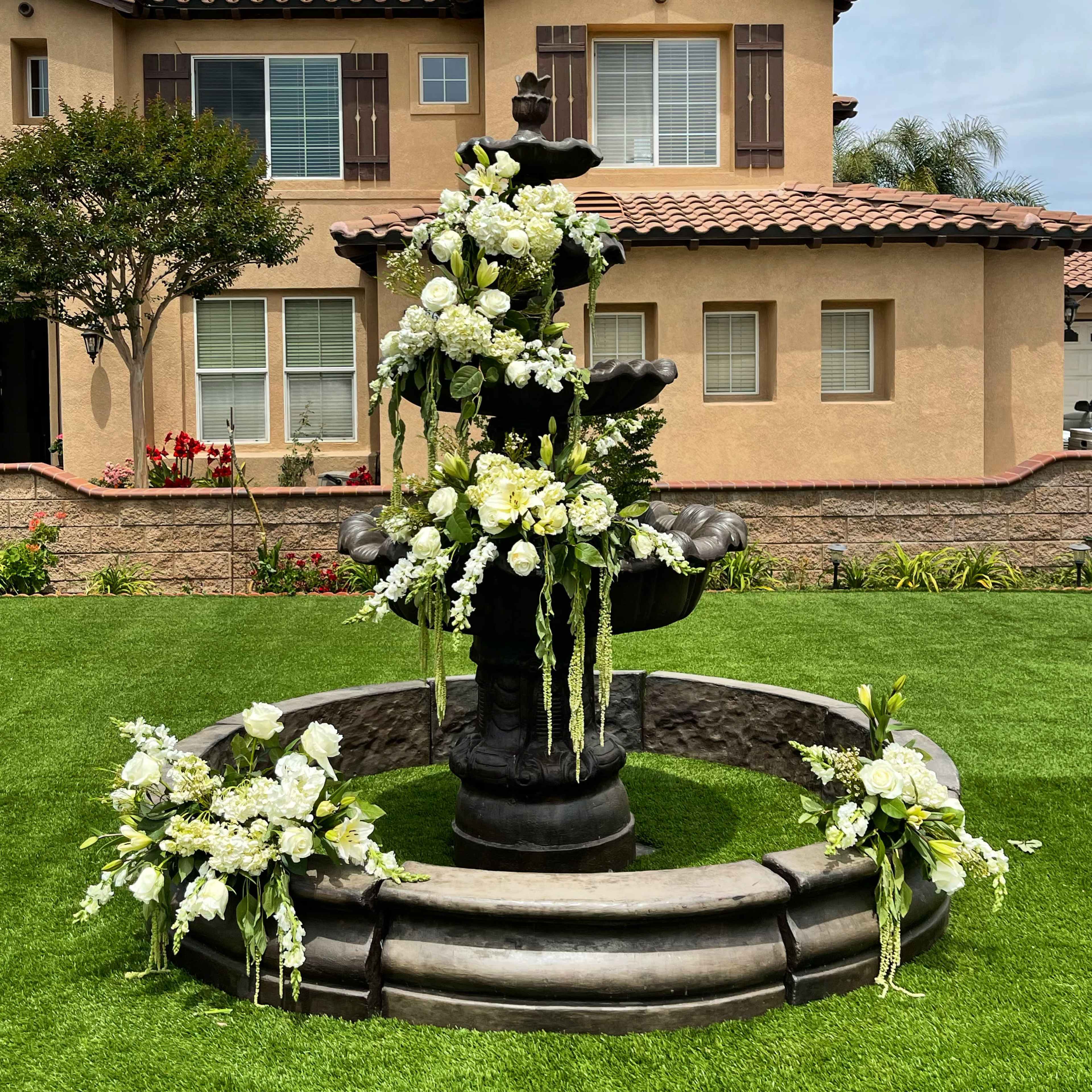 A black stone fountain adorned with white flowers is situated on a green lawn in front of a two-story house.