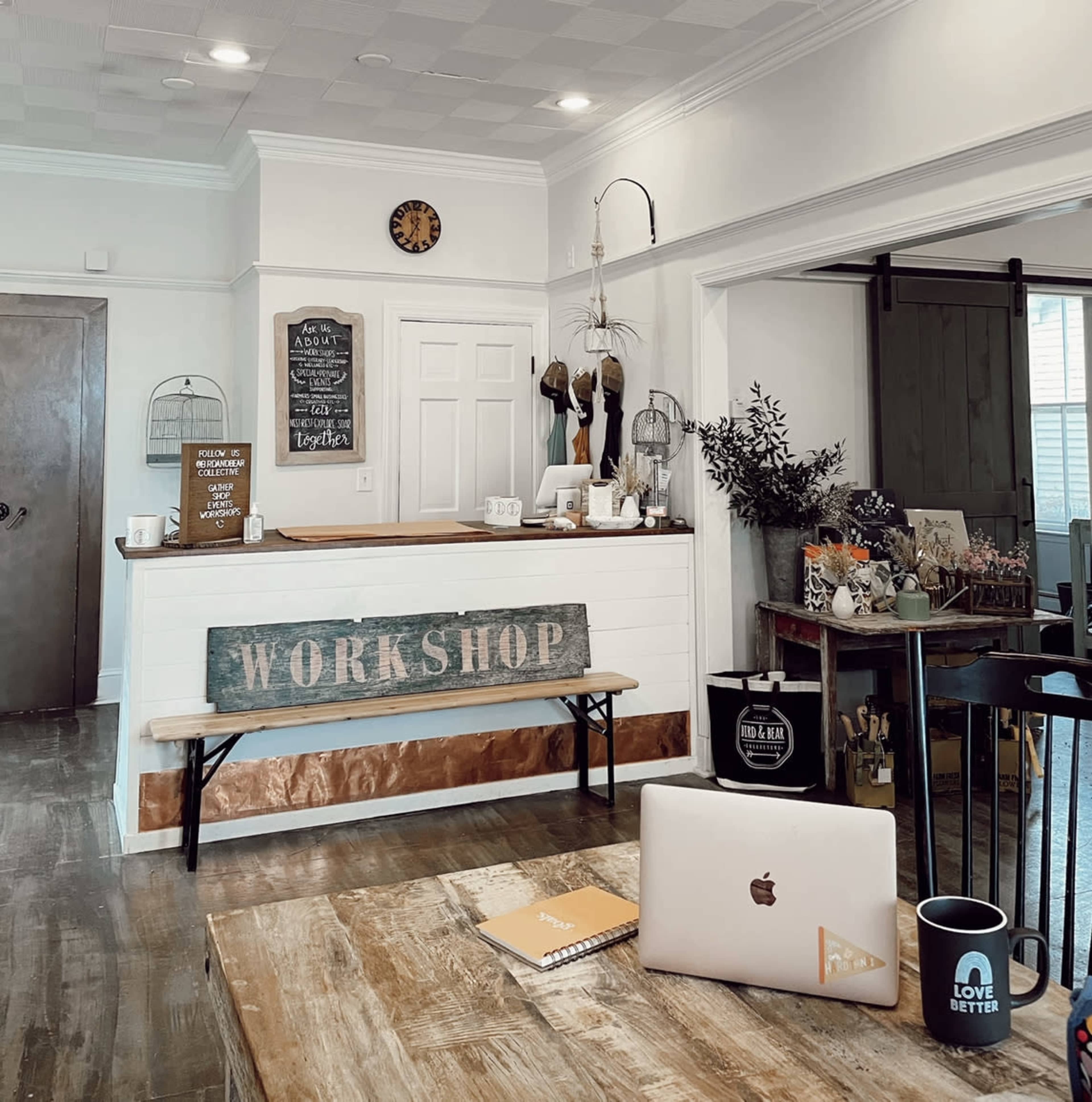 A cozy workshop reception area featuring a wooden counter, a sign that reads "WORKSHOP," and various decorative items like mugs, plants, and a laptop on a table.