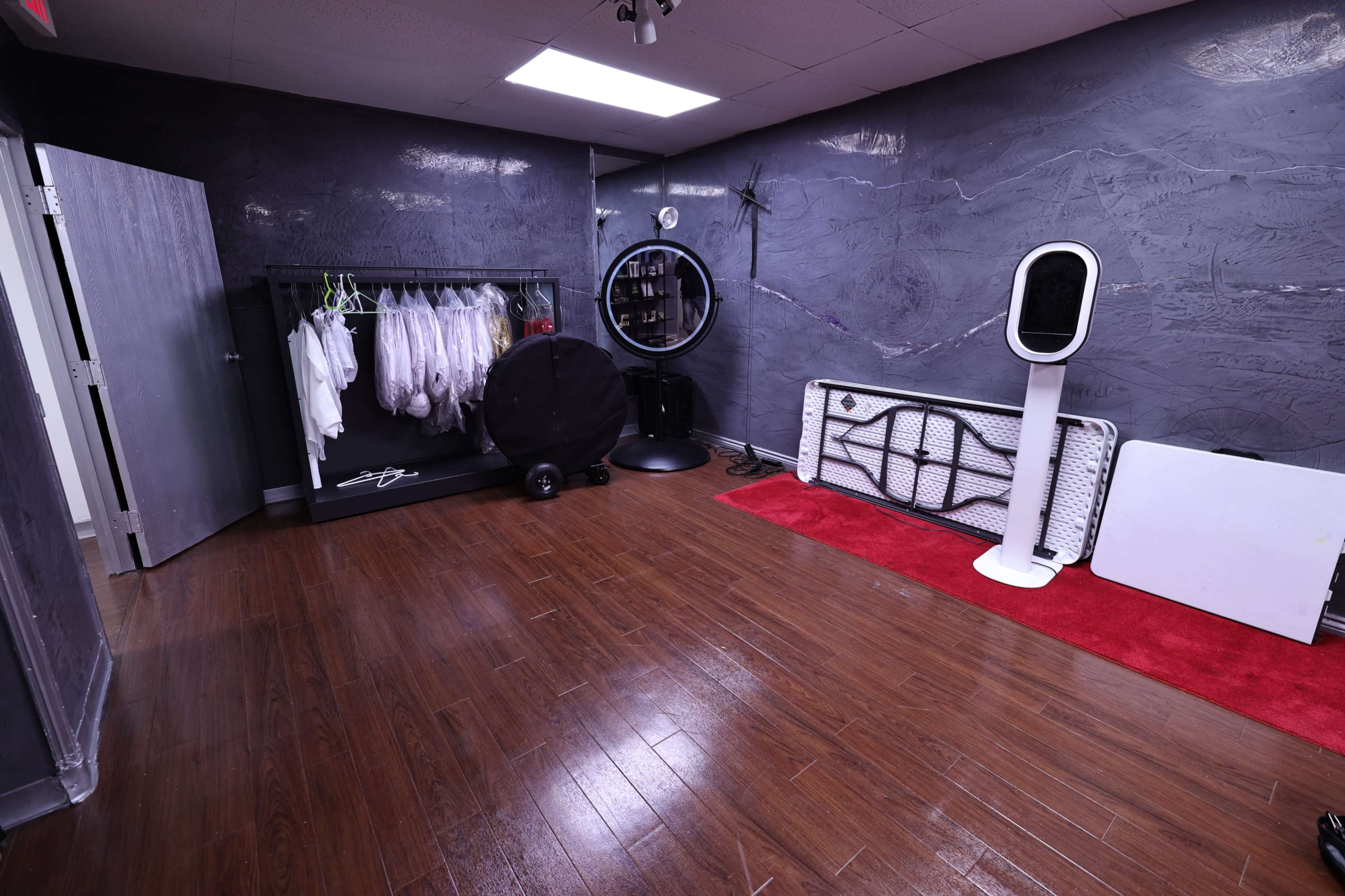A room with wooden flooring, featuring a clothing rack with white garments, a circular mirror, and various equipment along the walls.