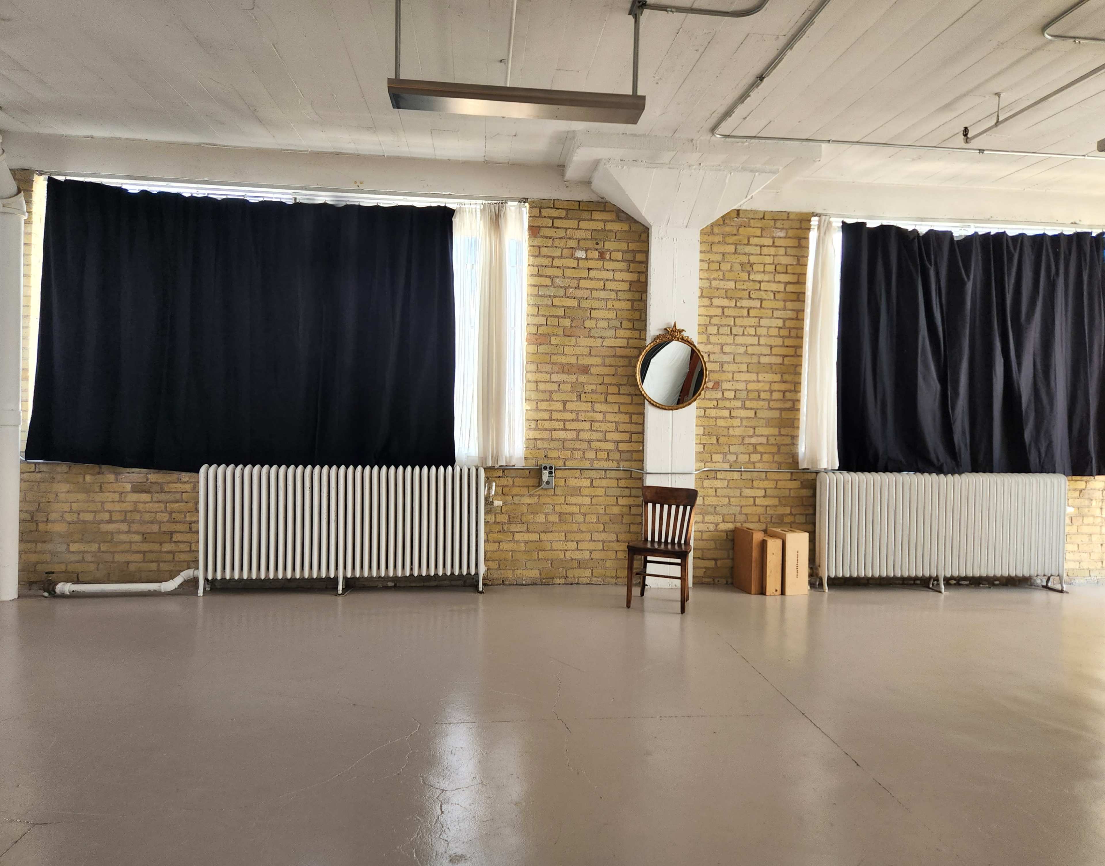 The image shows an empty room with exposed brick walls, a single chair, a mirror on the wall, and large windows covered by dark curtains.