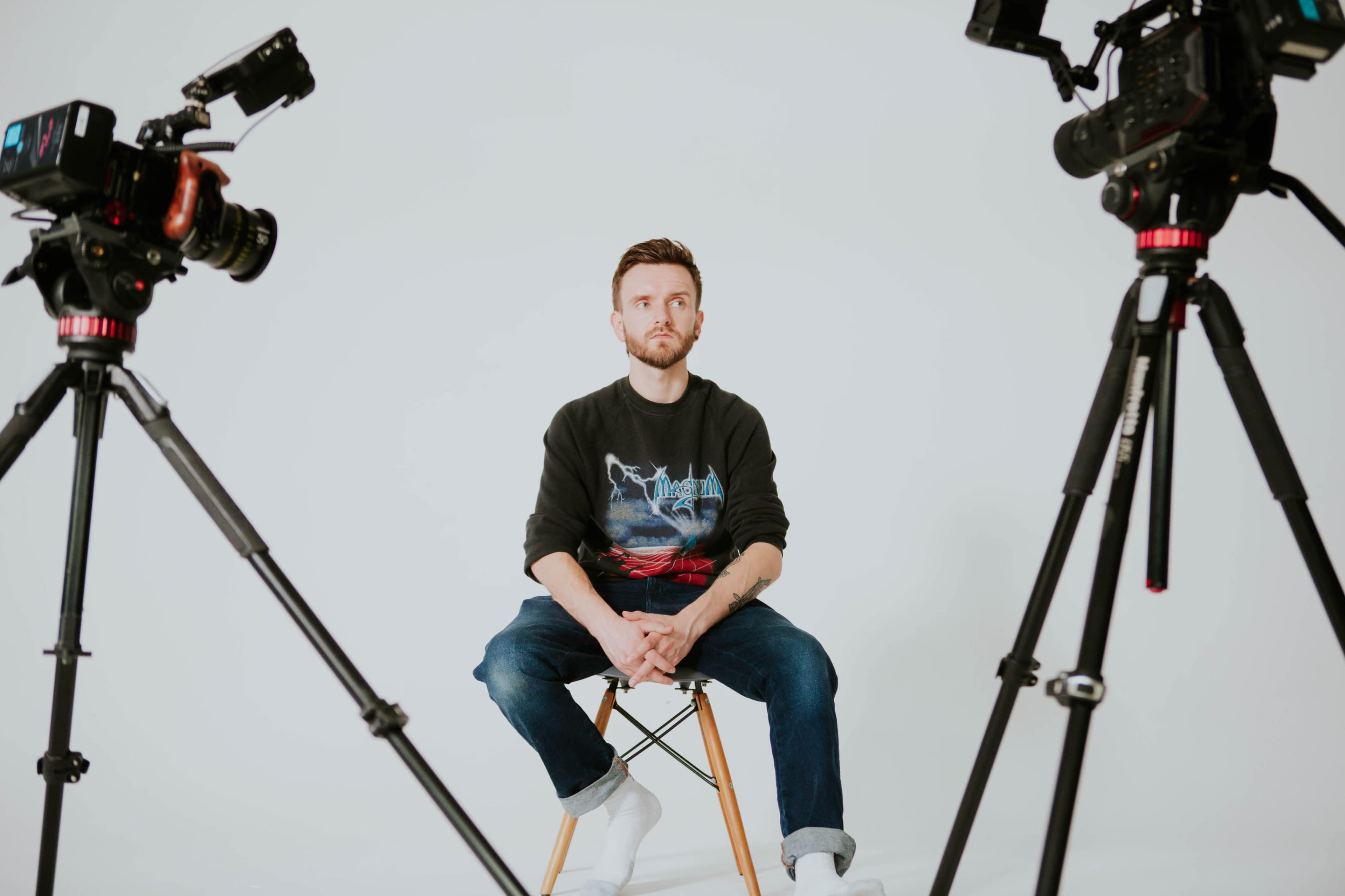 A man sits on a stool between two cameras on tripods against a plain white background.