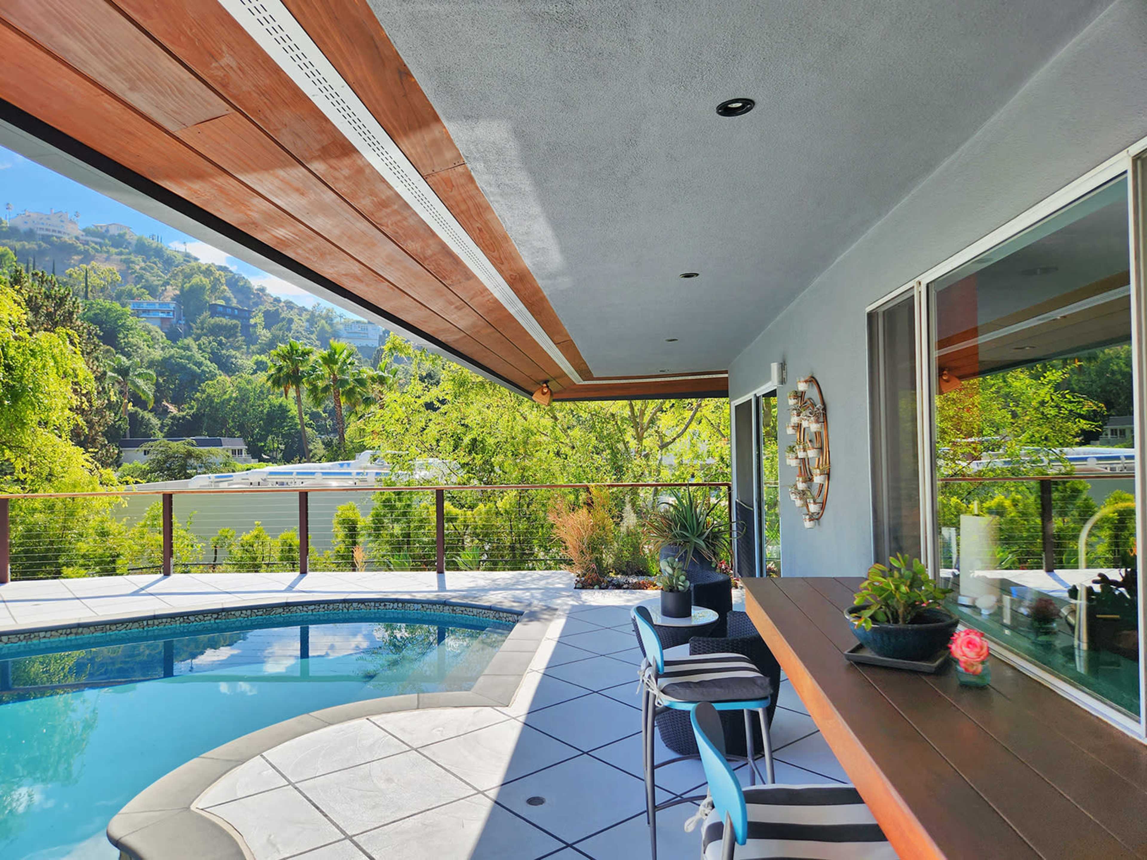 A modern outdoor patio area with a swimming pool, a bar counter, and lush greenery in the background.