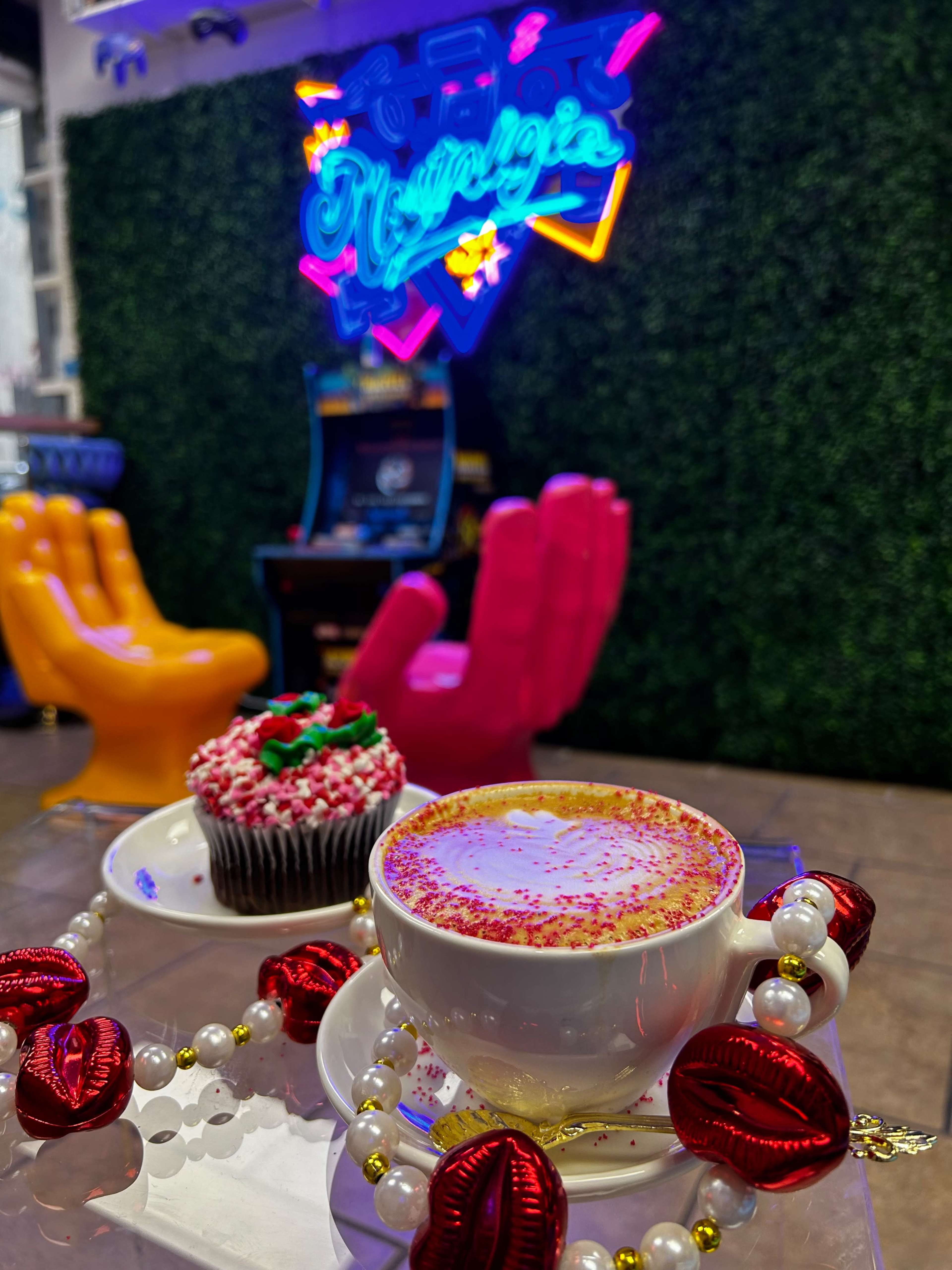 A decorated coffee cup with pink sprinkles sits on a red saucer next to a cupcake topped with frosting and a cherry, in front of a neon sign and vibrant chairs.