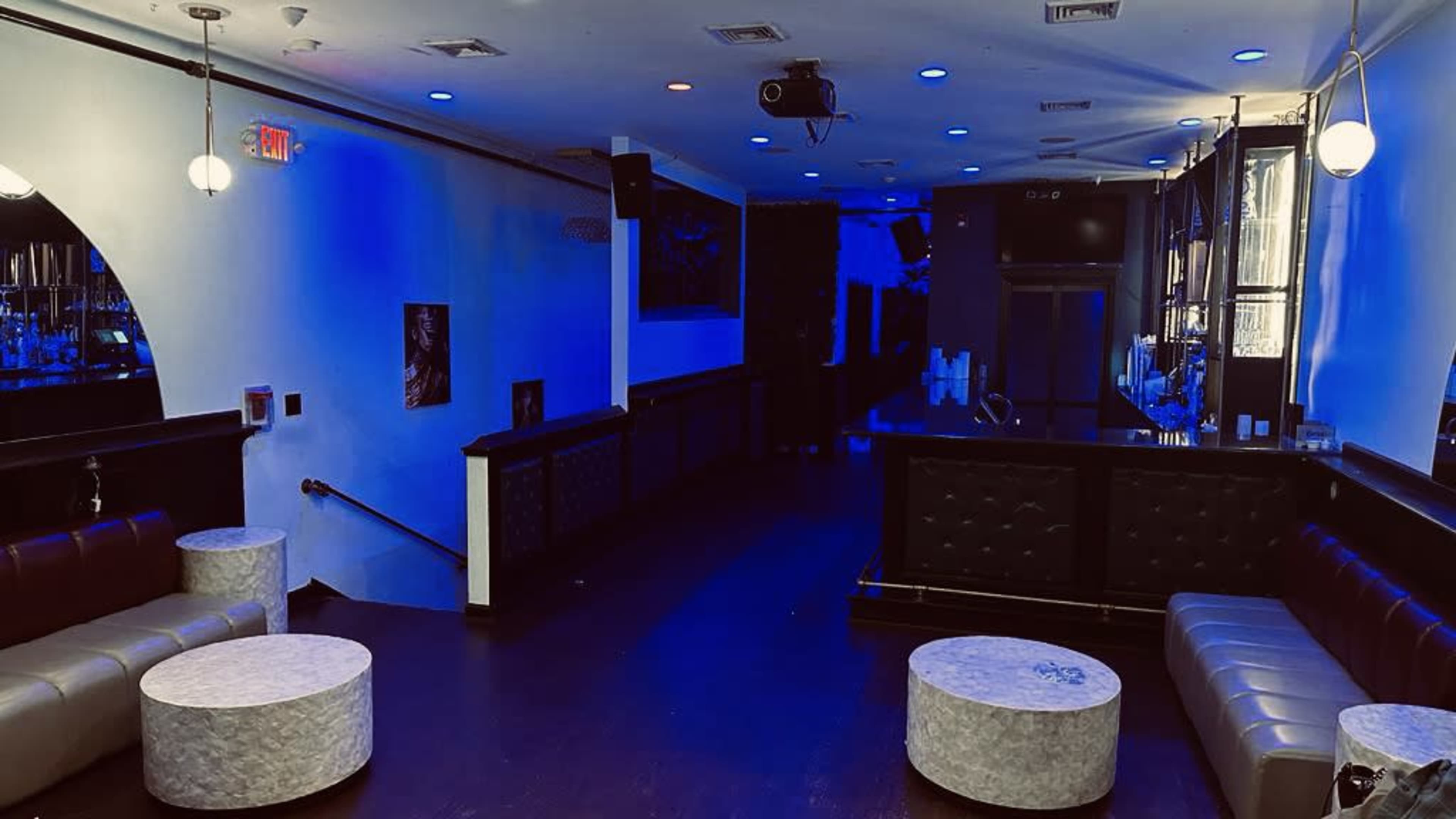 The image shows a dimly lit lounge with blue lighting, featuring a bar area and several round tables surrounded by seating along the walls.