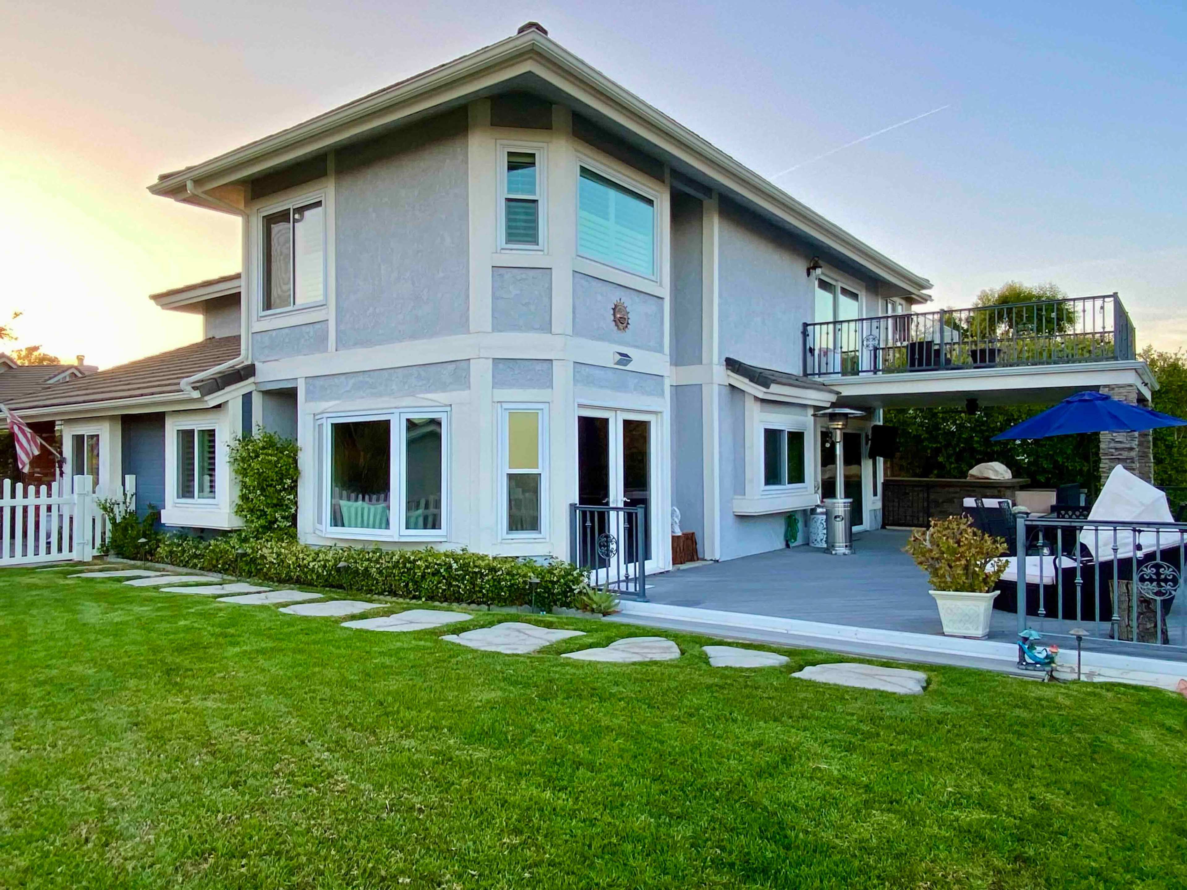 A modern two-story house features large windows and a deck overlooking a landscaped yard with a stone path.