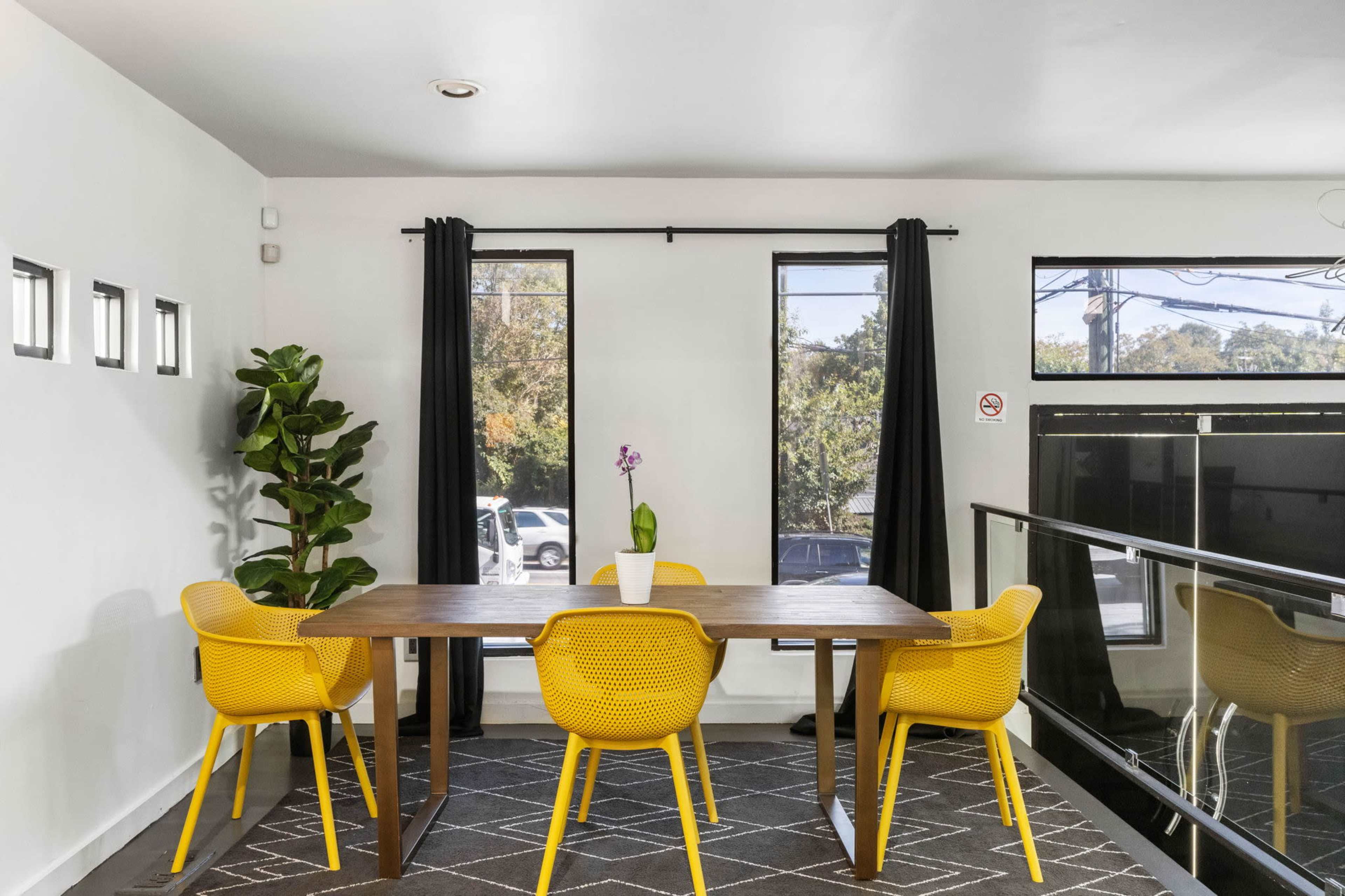 A dining area features a wooden table surrounded by four yellow chairs, with large windows and a potted plant in the corner.