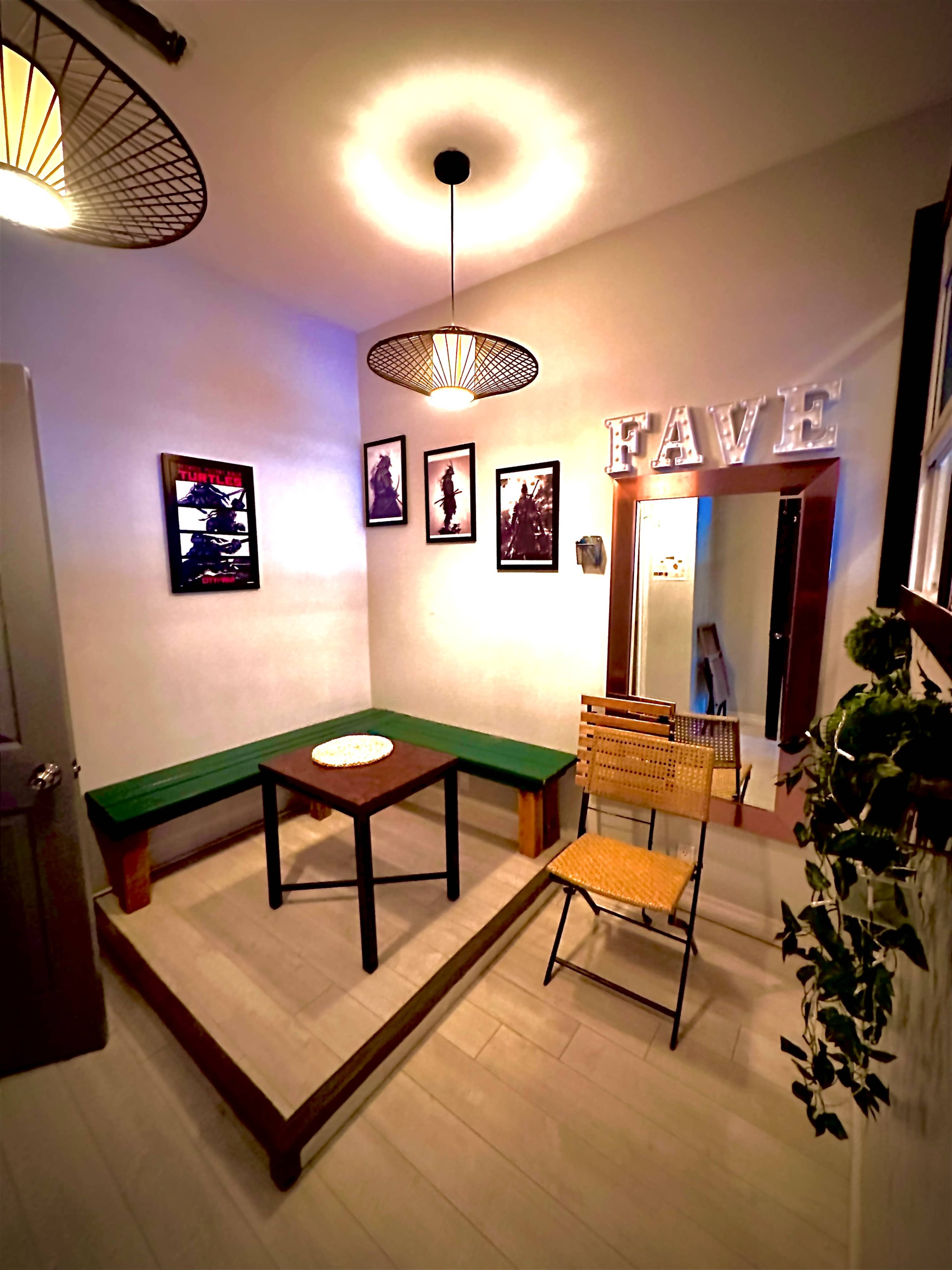 The image shows a small, well-lit room featuring a green bench, a wooden table, a folding chair, several framed pictures on the walls, and a mirror.