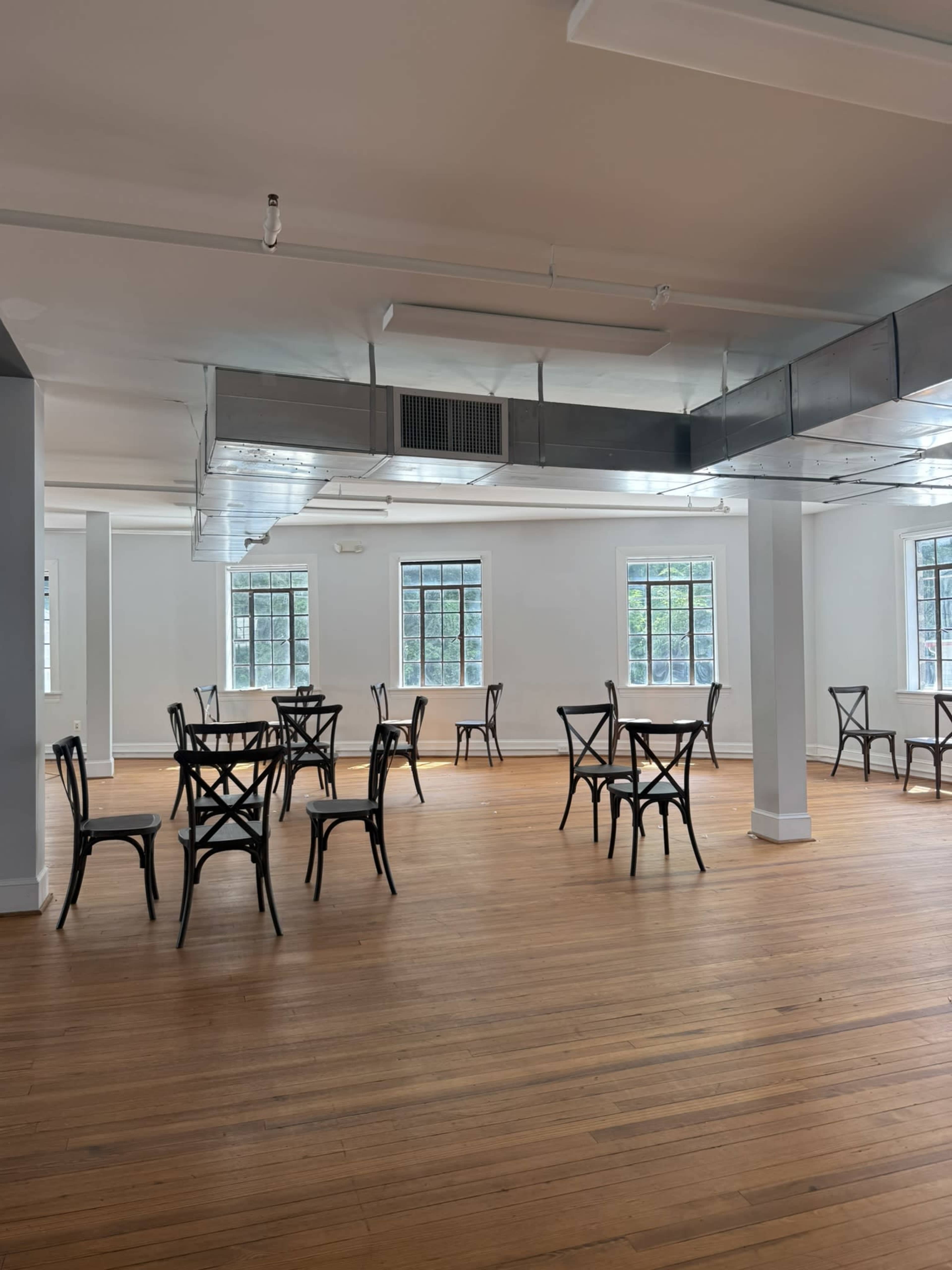The image shows a spacious, empty room with wooden flooring and several black tables and chairs arranged throughout, set against large windows.