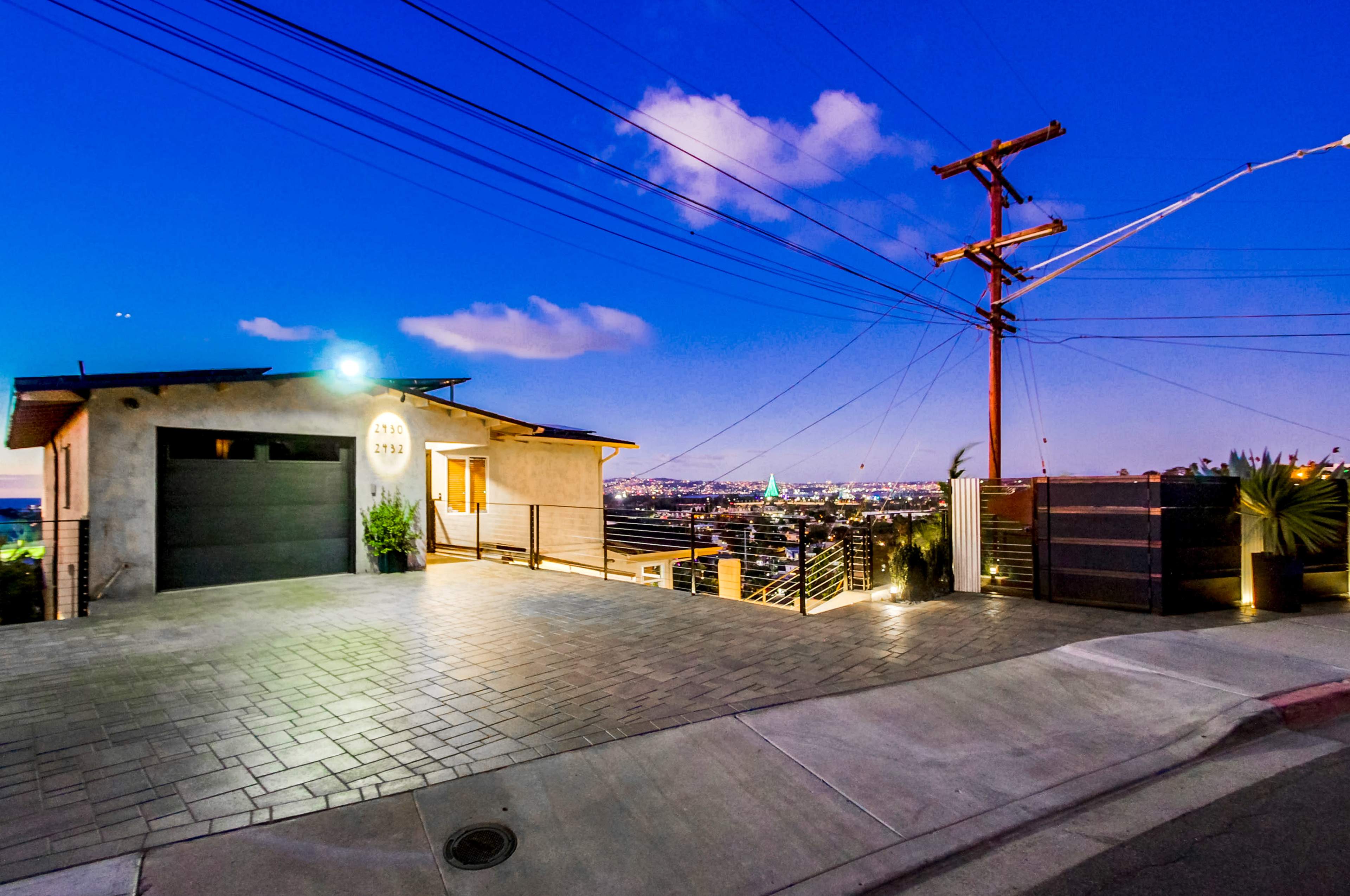 A modern house with a dark garage and a paved driveway overlooks a city skyline at dusk, framed by utility poles and power lines.