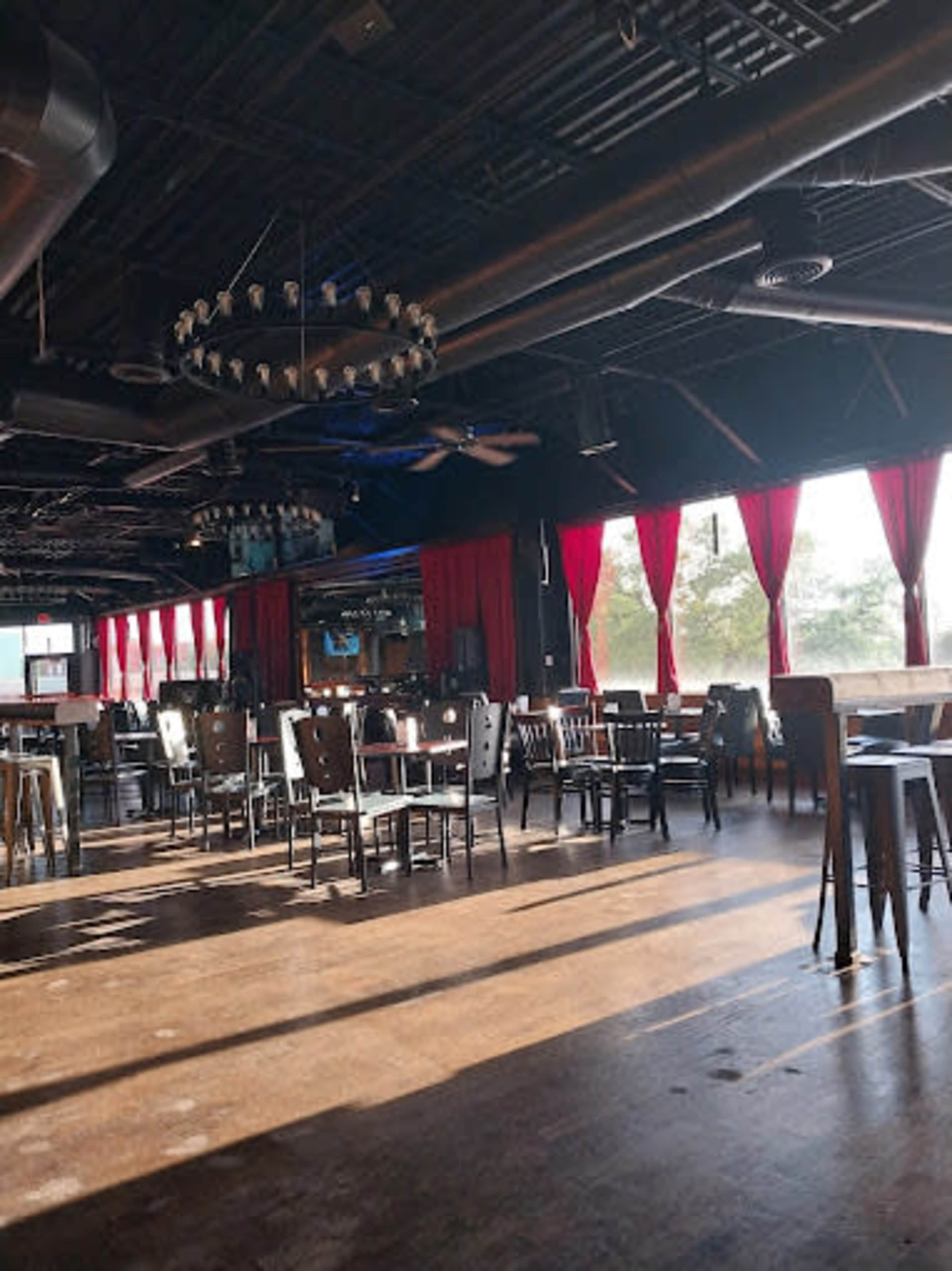 The interior of a spacious restaurant features wooden tables and chairs, large windows with red curtains, and industrial-style lighting fixtures.