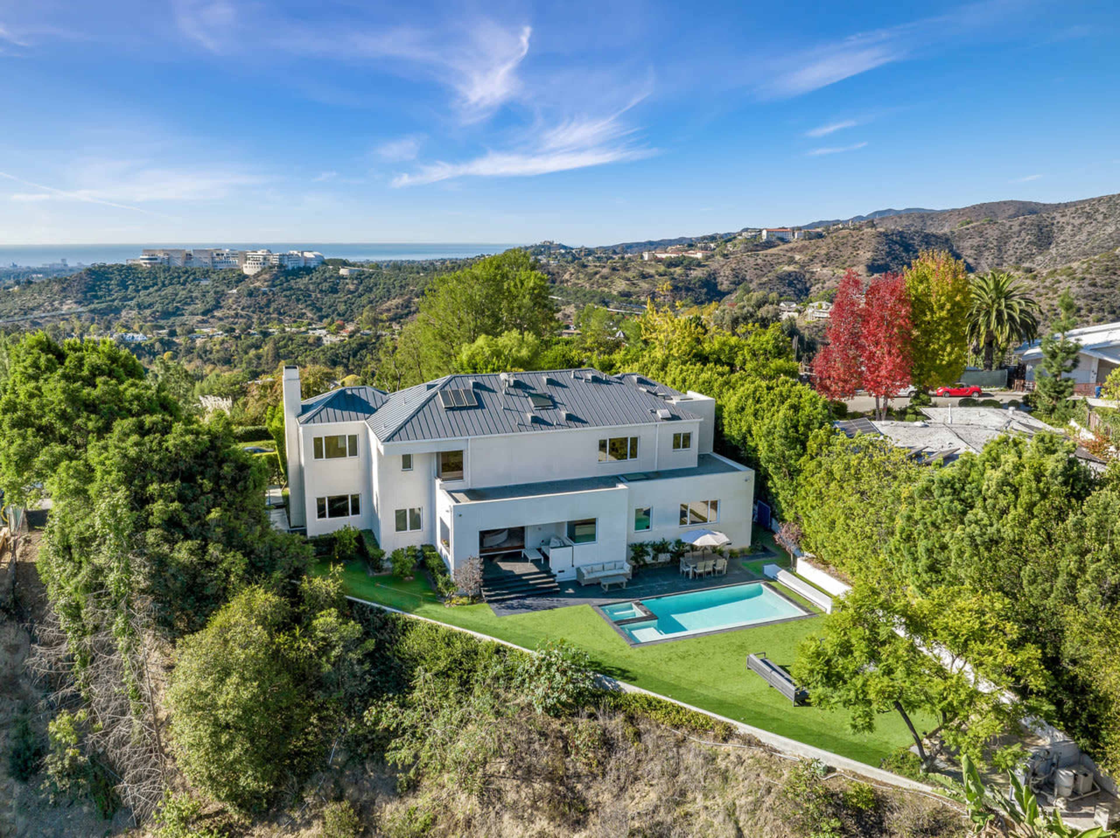 A modern two-story house with a pool is set on a hillside, surrounded by greenery and featuring a view of the distant hills.