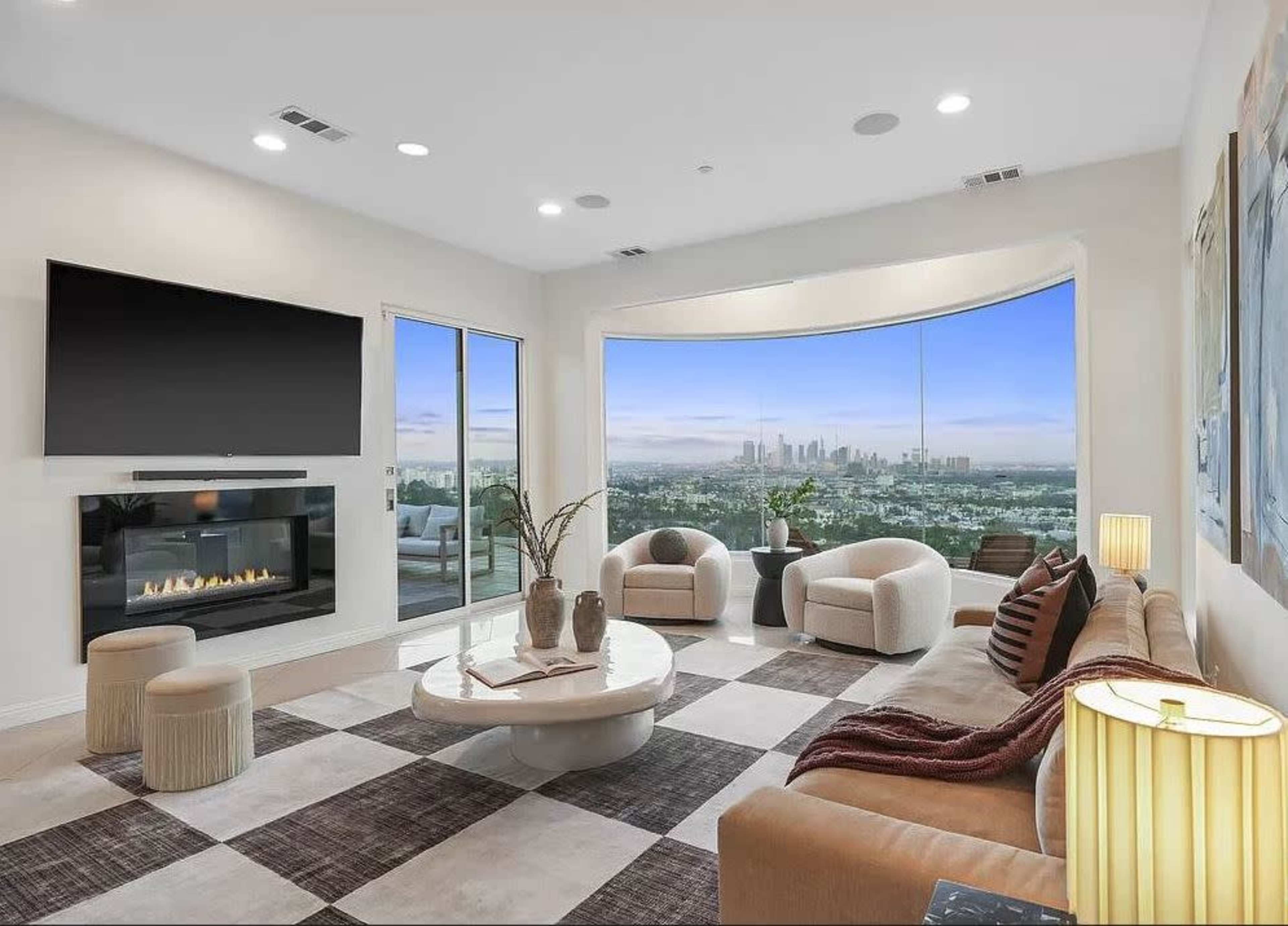 The spacious living room features a large window with a view of the city skyline, a modern fireplace, and contemporary furniture arranged on a checkered floor.