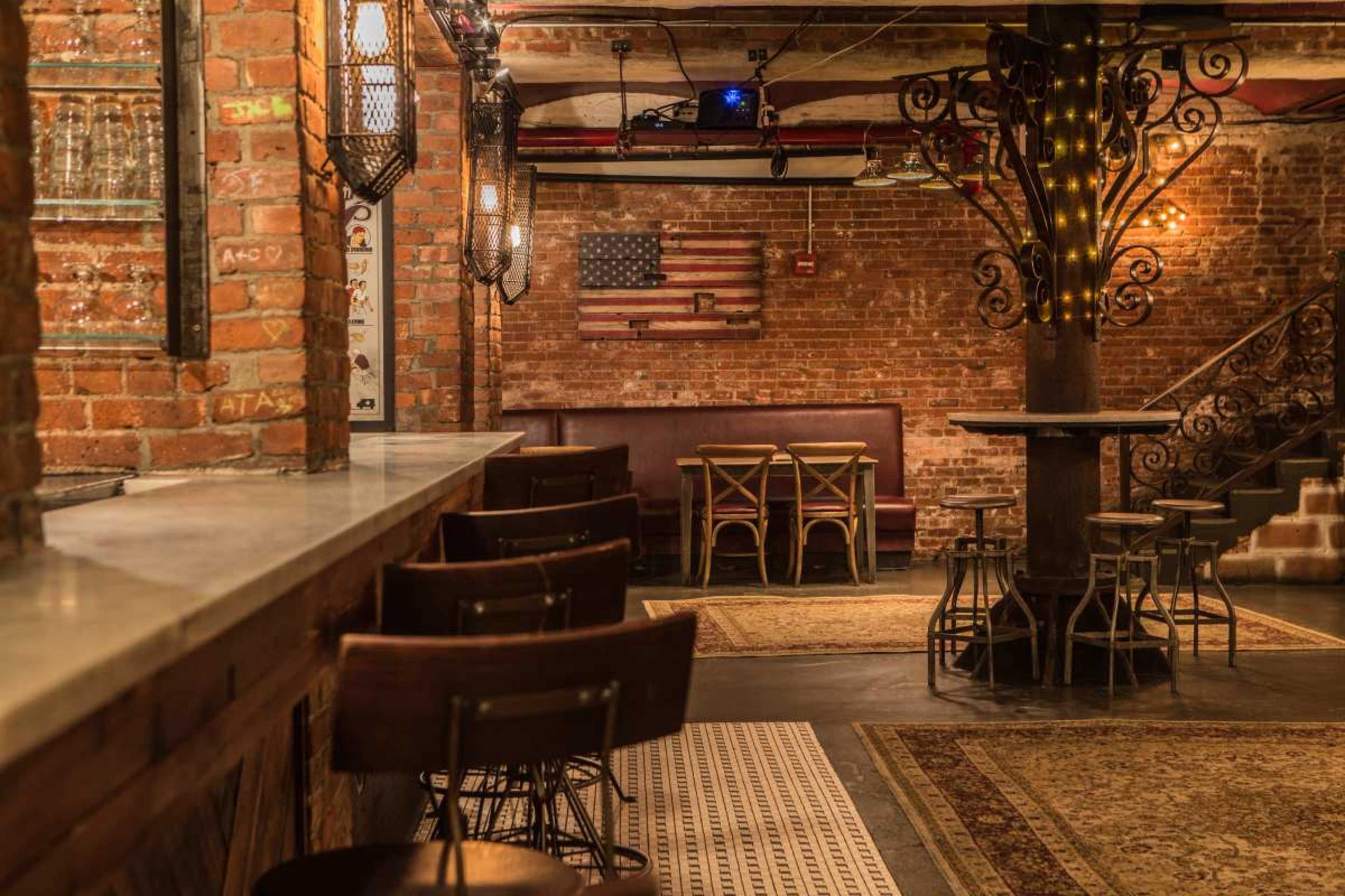 The image shows a cozy bar area with a brick wall, wooden furniture, and a staircase leading to an upper level, featuring an American flag and ambient lighting.