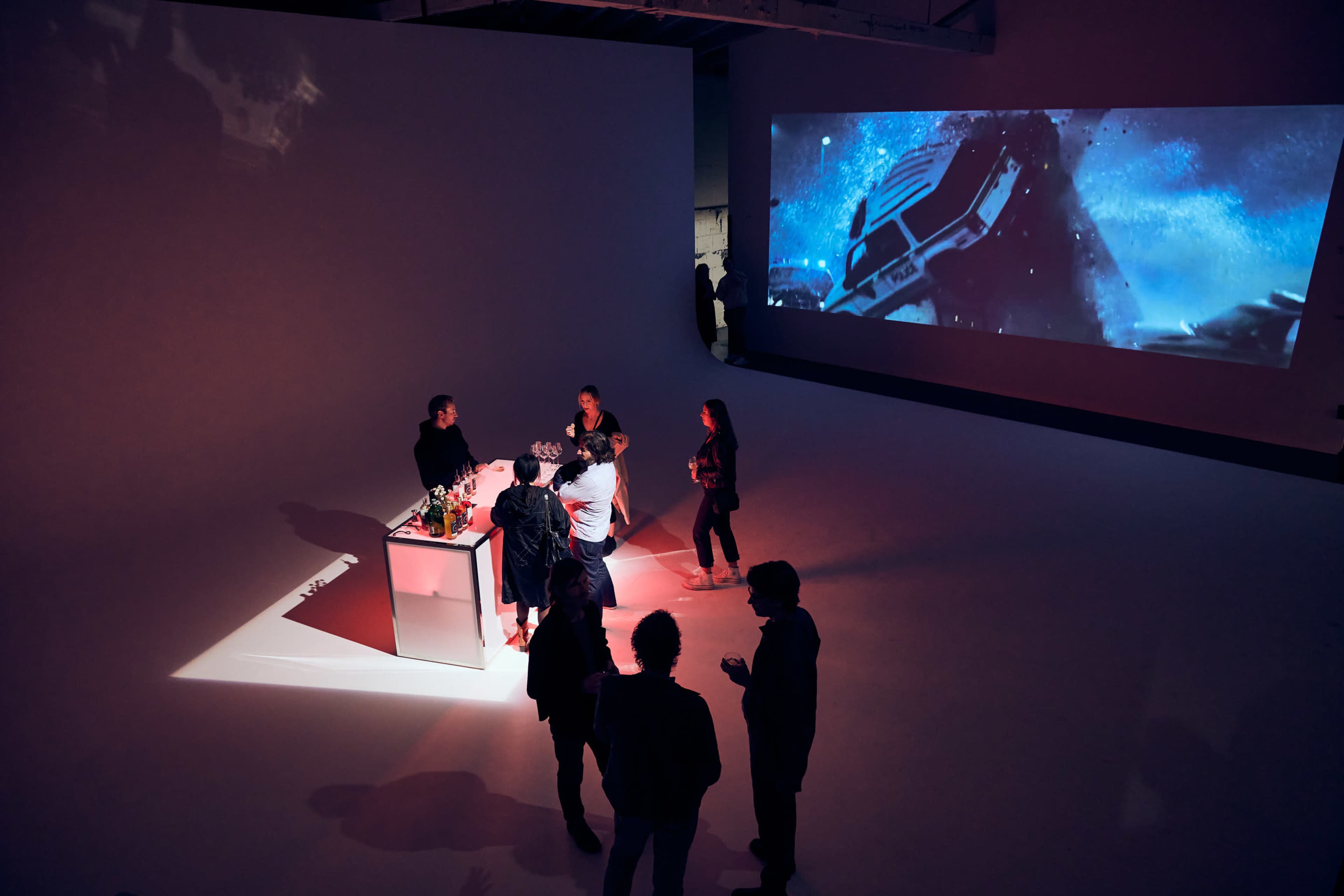 A group of people gathers around a bar in a dimly lit space, with a large video projection on the wall displaying a dramatic scene.