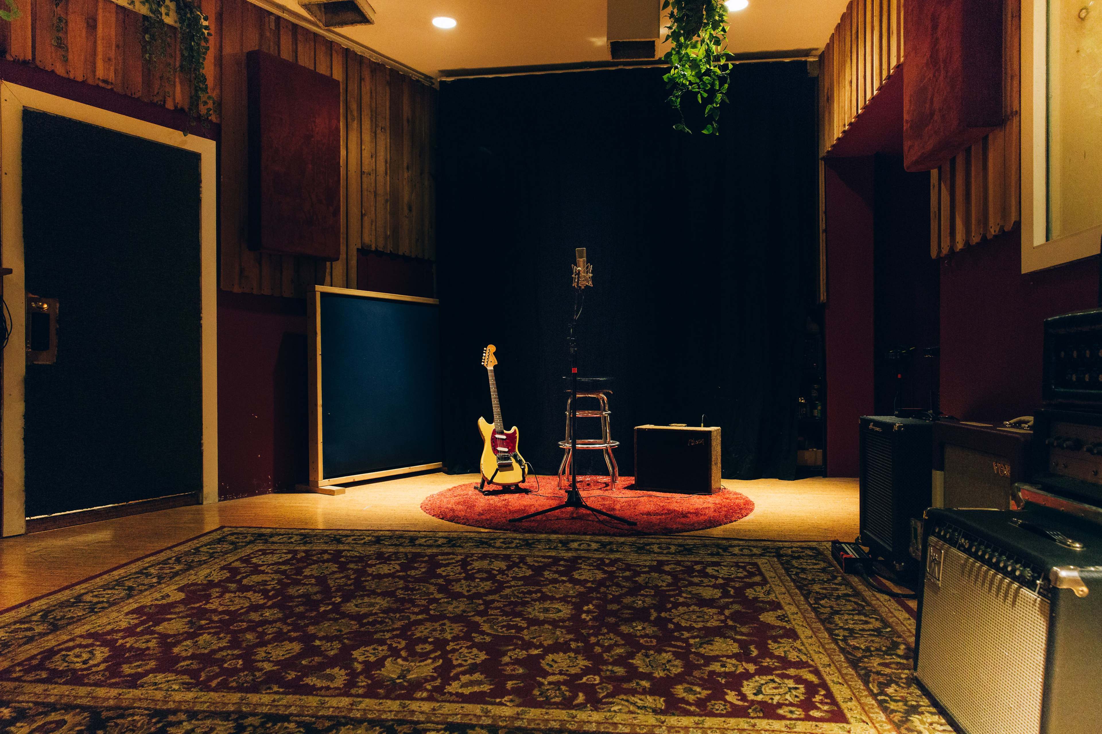 A microphone on a stool is positioned in front of a guitar and amplifier in a dimly lit recording studio.
