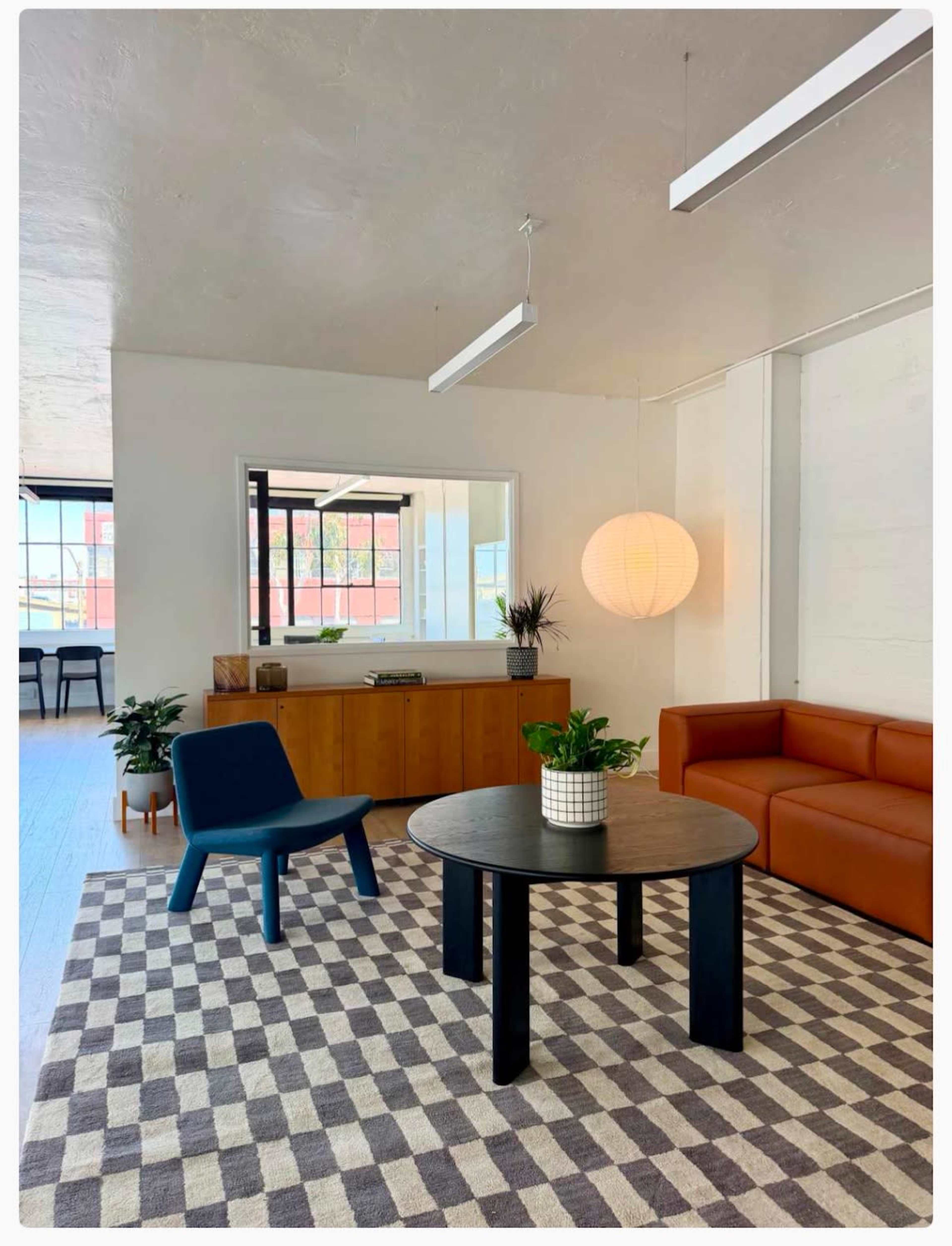 The image shows a modern interior space featuring a brown leather sofa, a round black table, a blue chair, a patterned rug, and large windows letting in natural light.