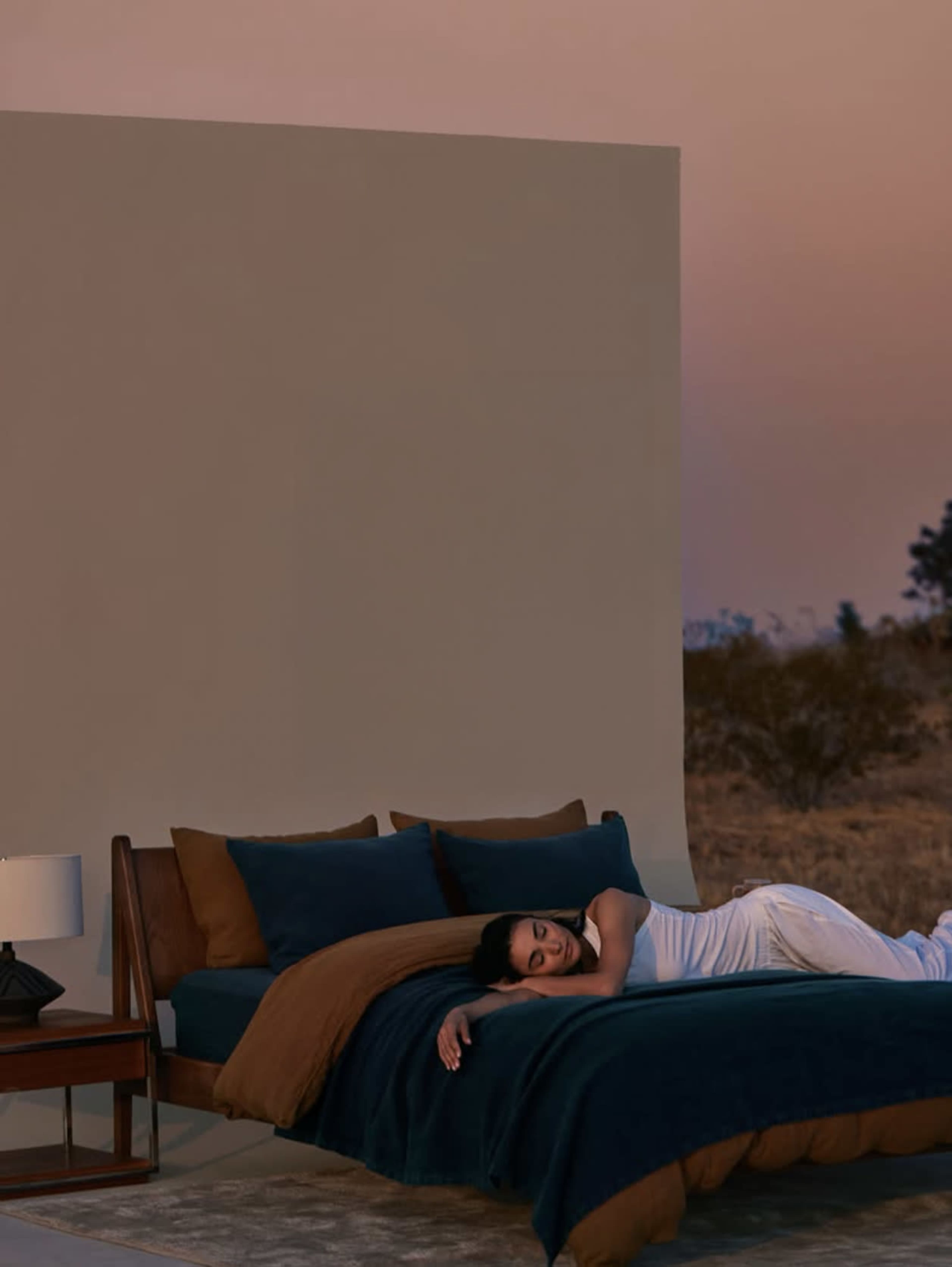 A woman sleeps on a bed with blue and brown bedding set against a simple backdrop during sunset.