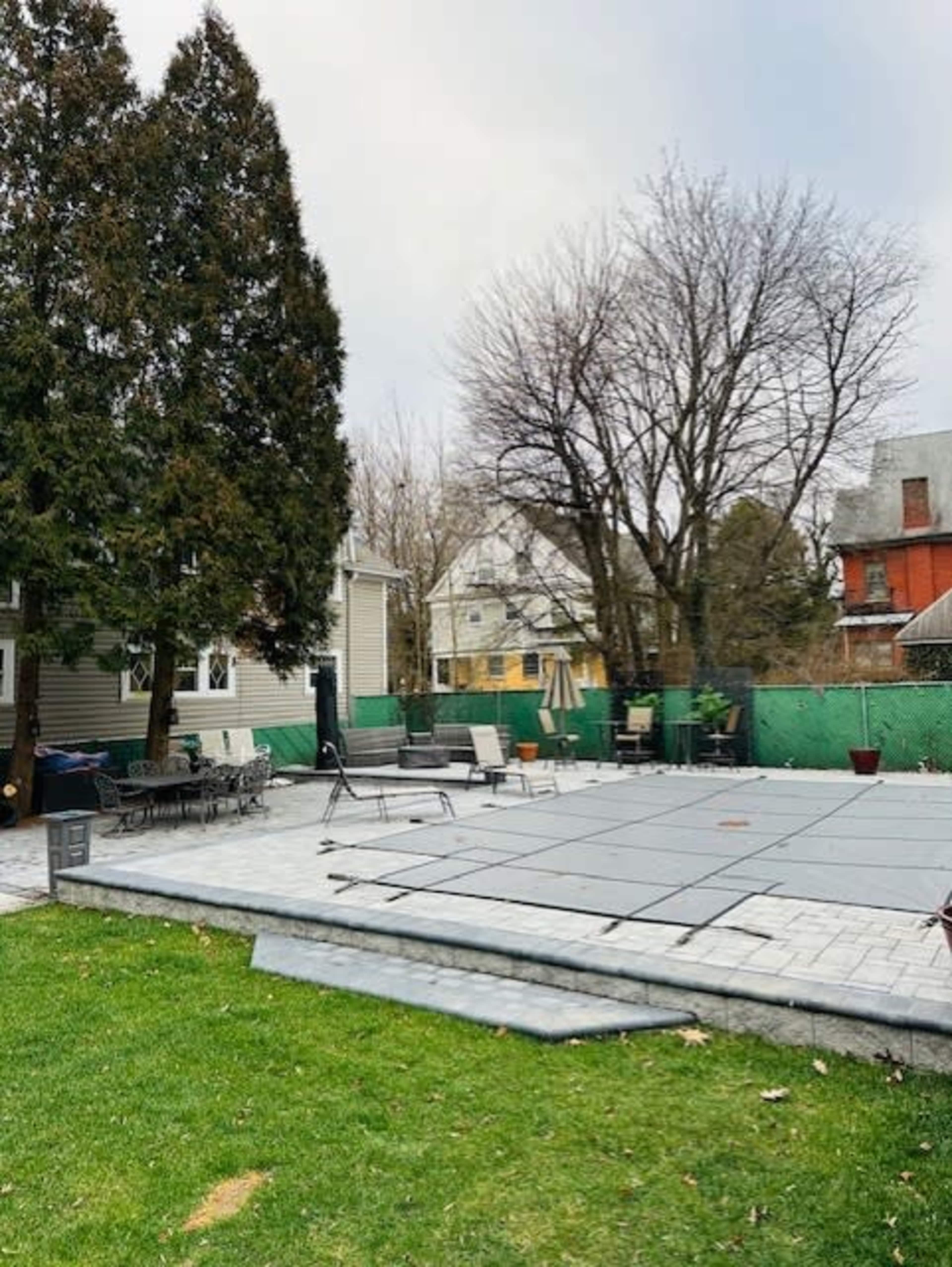 A backyard scene features a swimming pool area covered with a safety cover, surrounded by patio furniture and trees.