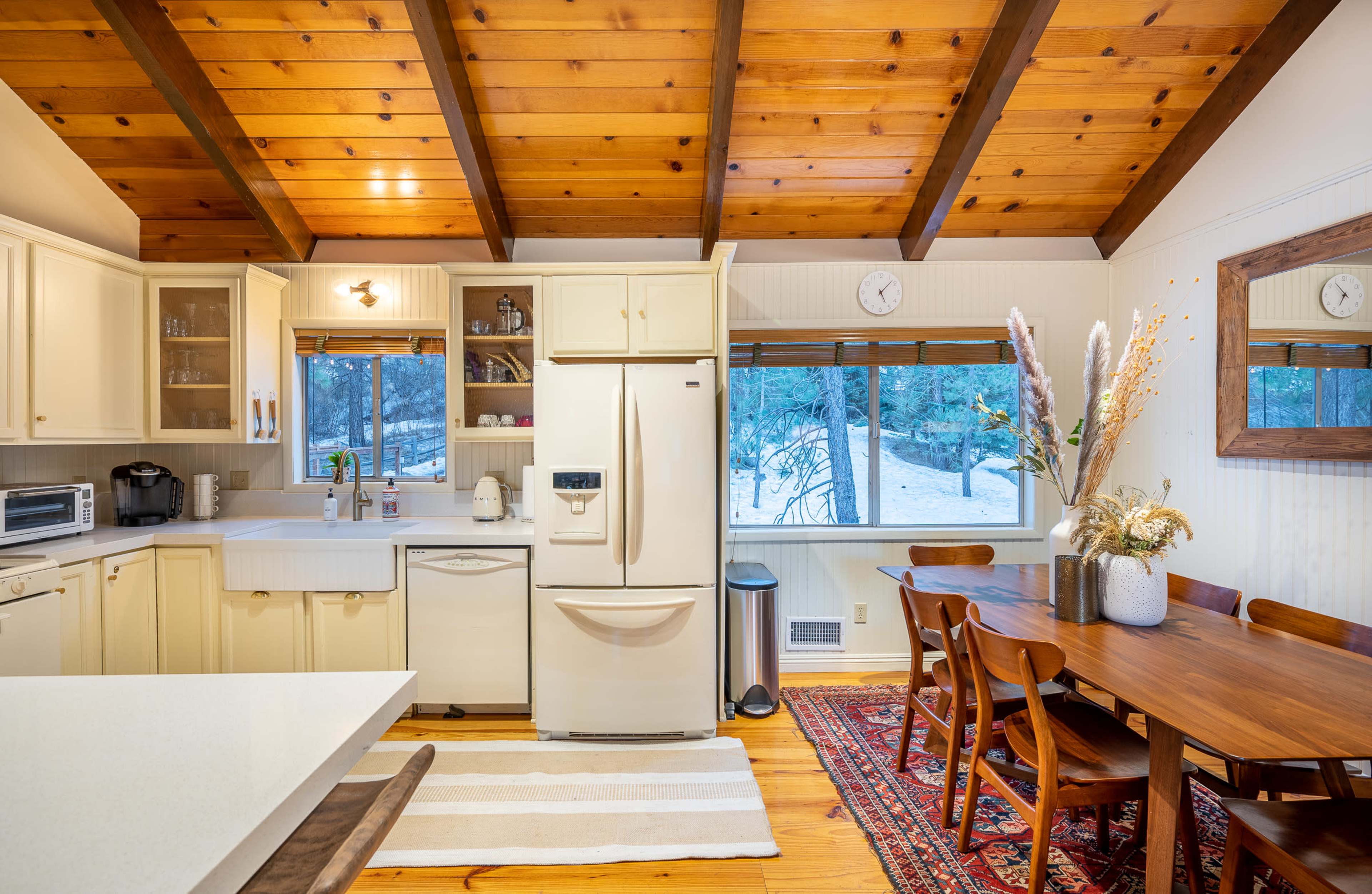 The image shows a kitchen with wooden beams, light-colored cabinets, and a dining table, featuring a view of a snowy outdoor scene through a window.