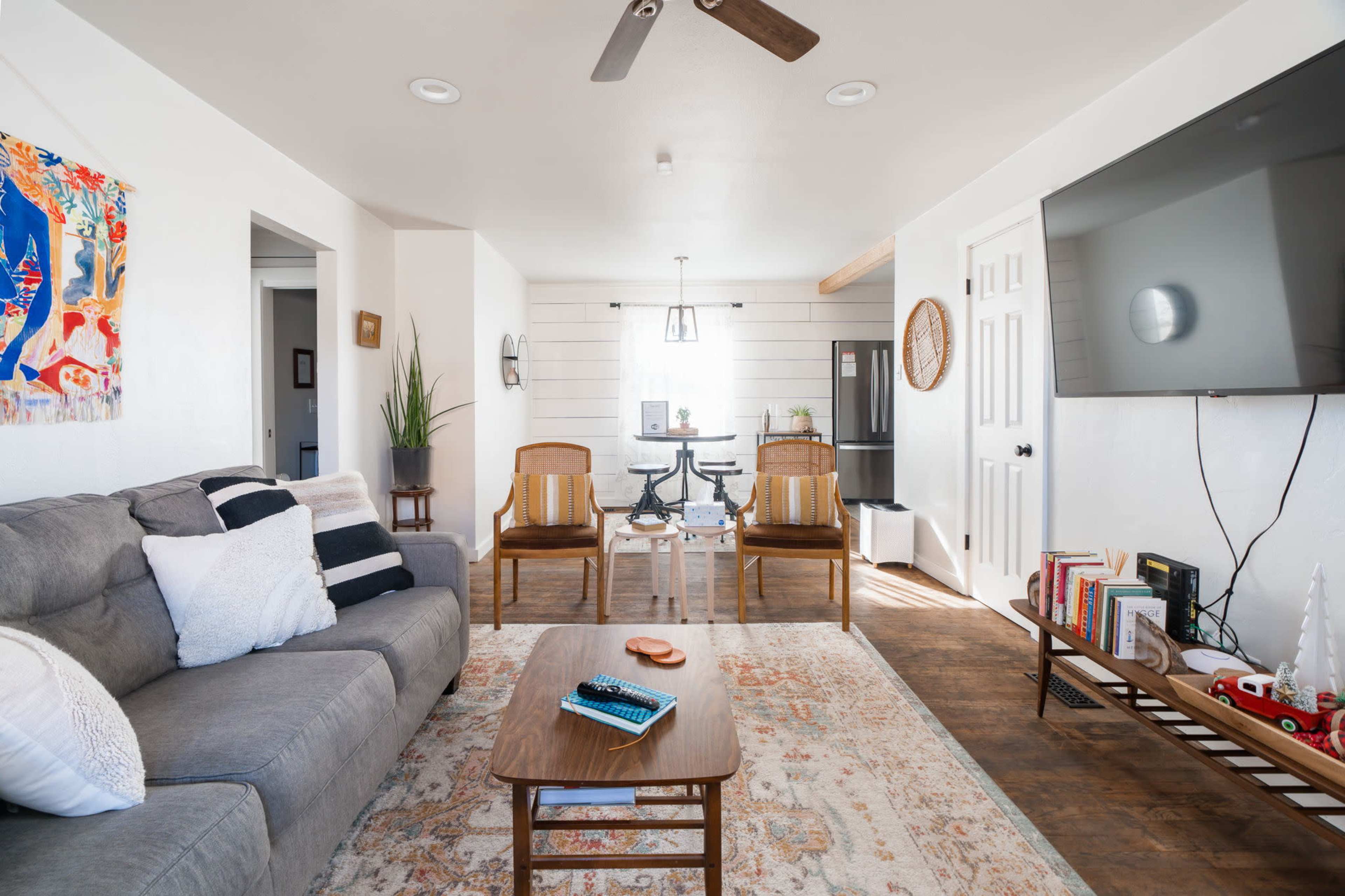 A cozy living room features a gray sofa, an area rug, a wooden coffee table, and a dining area with a glass table and chairs, all illuminated by natural light.