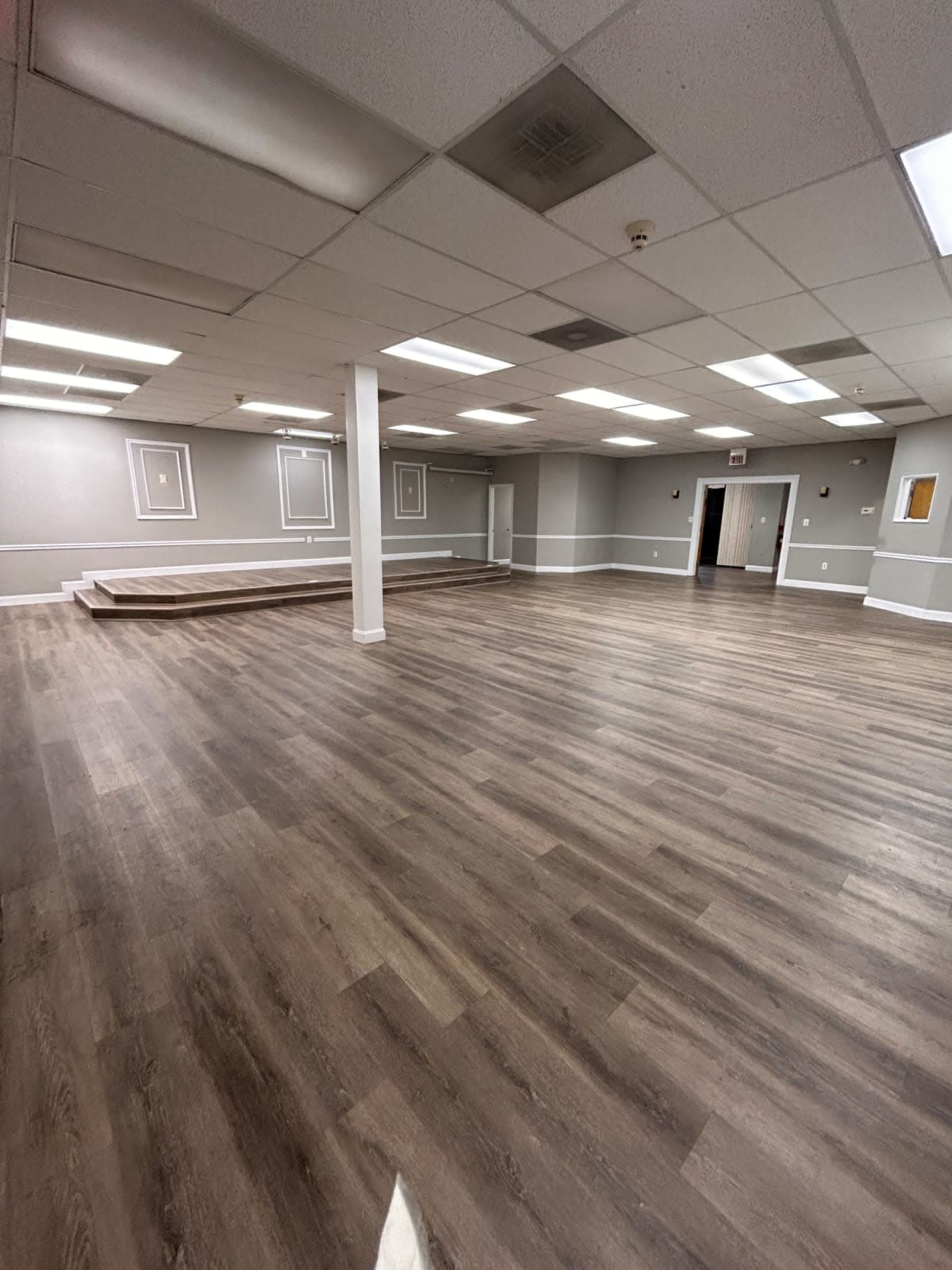 The image shows a spacious, empty room with wooden flooring, white walls, and a raised platform at one end.