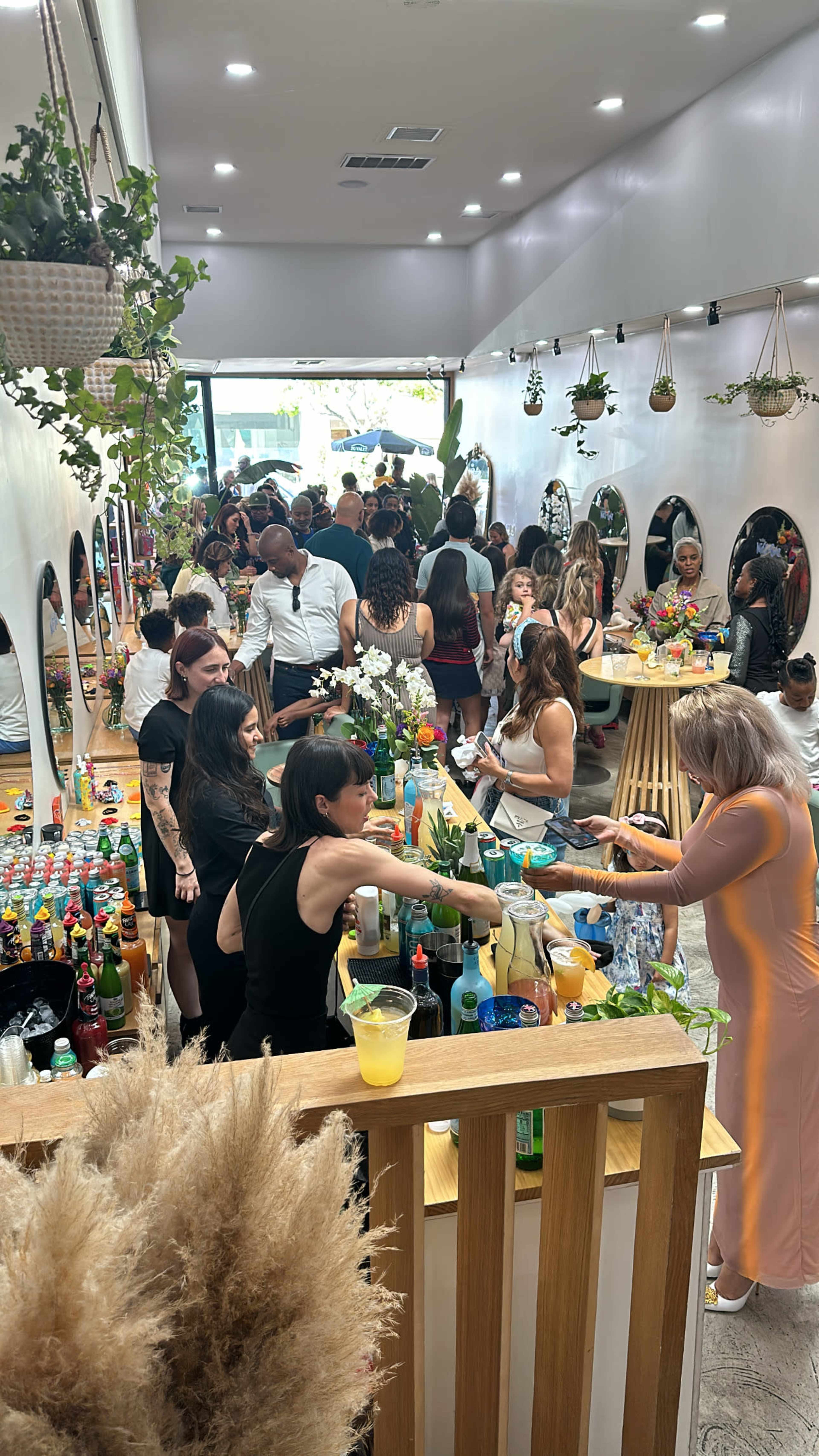 The image shows a busy bar scene with a crowd of people socializing and preparing drinks at a long counter adorned with colorful bottles and plants.