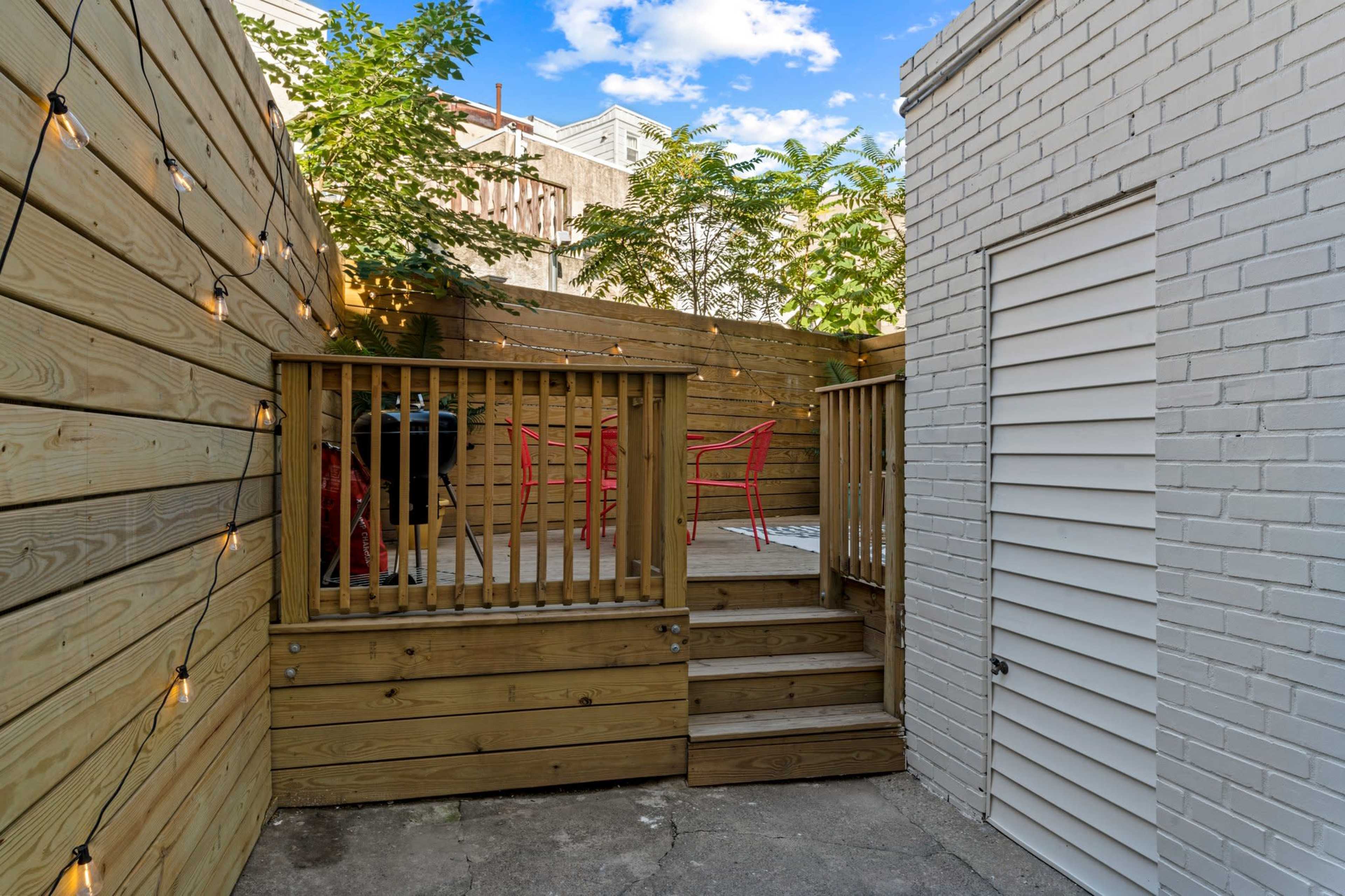 The image shows a small outdoor patio area with wooden decking, red chairs, and surrounding greenery, enclosed by wooden fences and a white brick wall.