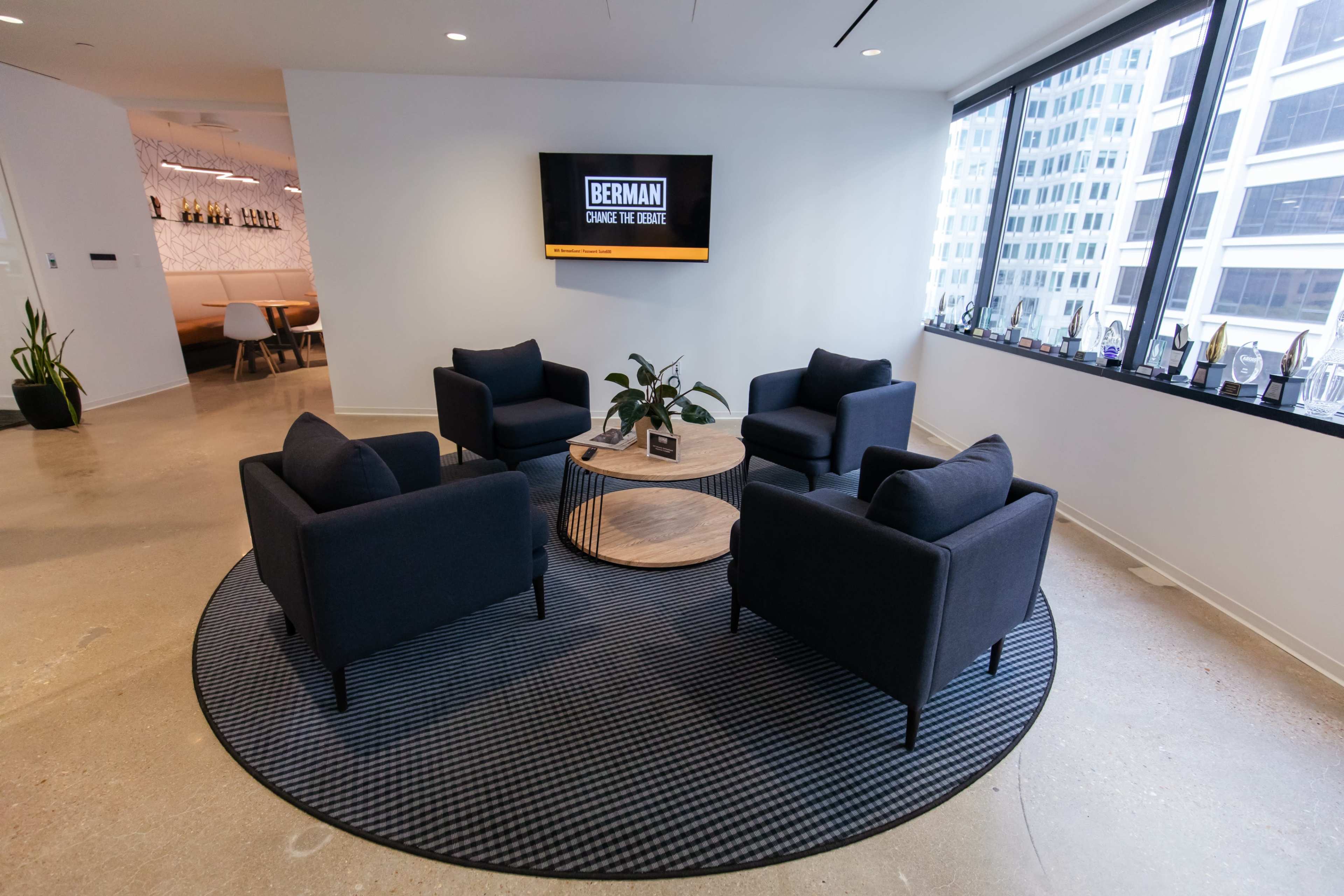 A modern office lounge area features four black chairs arranged around a round wooden coffee table on a patterned rug, with a television mounted on the wall displaying the name "BERMAN."