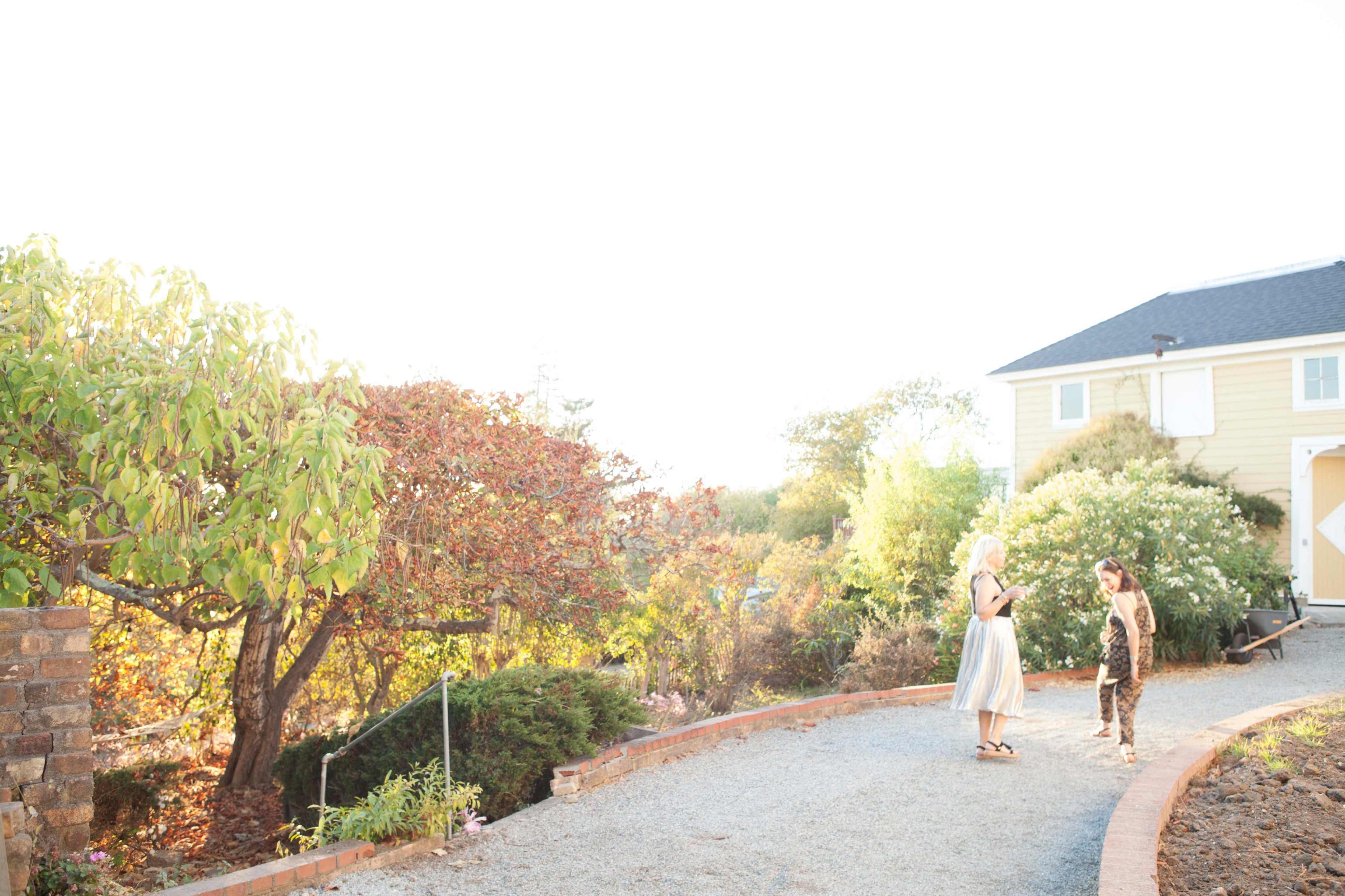 A woman and a child are standing on a gravel path in front of a house with autumn foliage in the background.