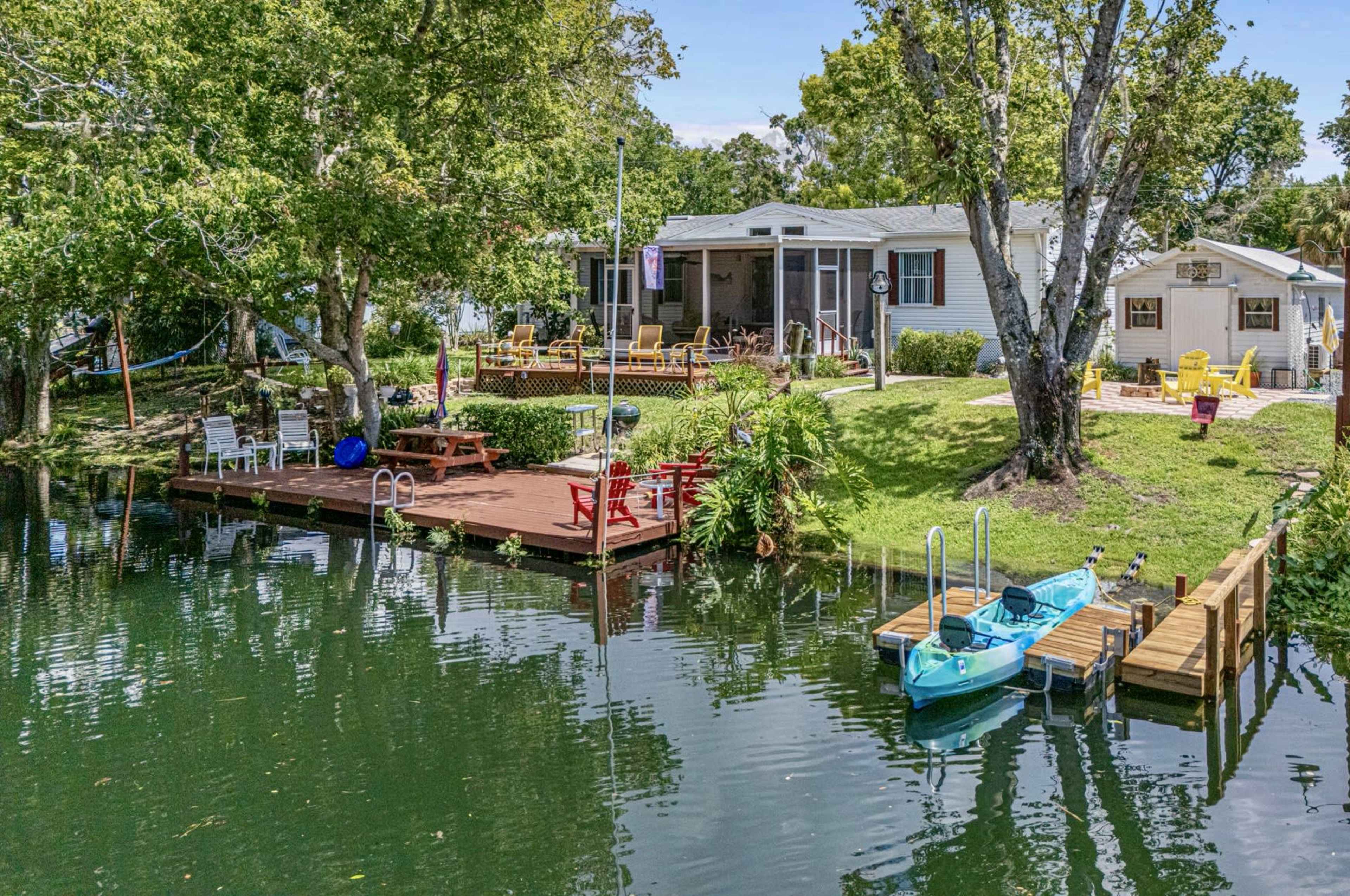 A waterfront property features a wooden deck with seating, two kayaks, and lush greenery surrounding a home, all adjacent to calm water.