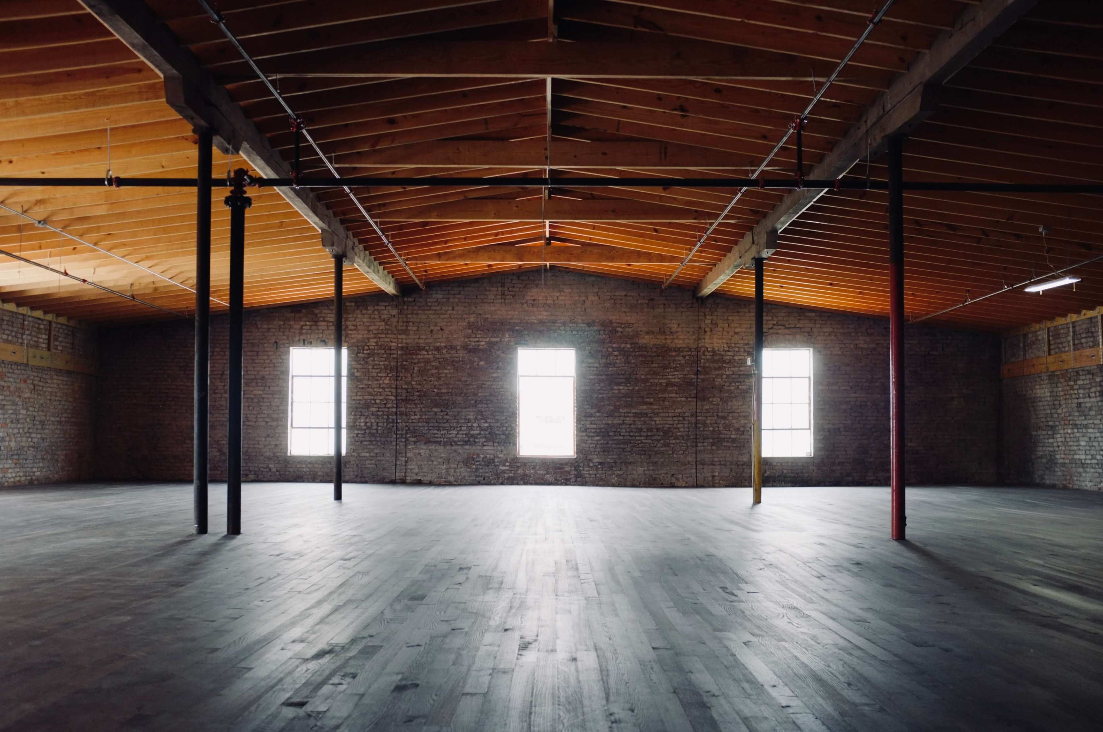 The image shows an empty industrial space with wooden beams, a polished floor, and large windows allowing natural light to enter.
