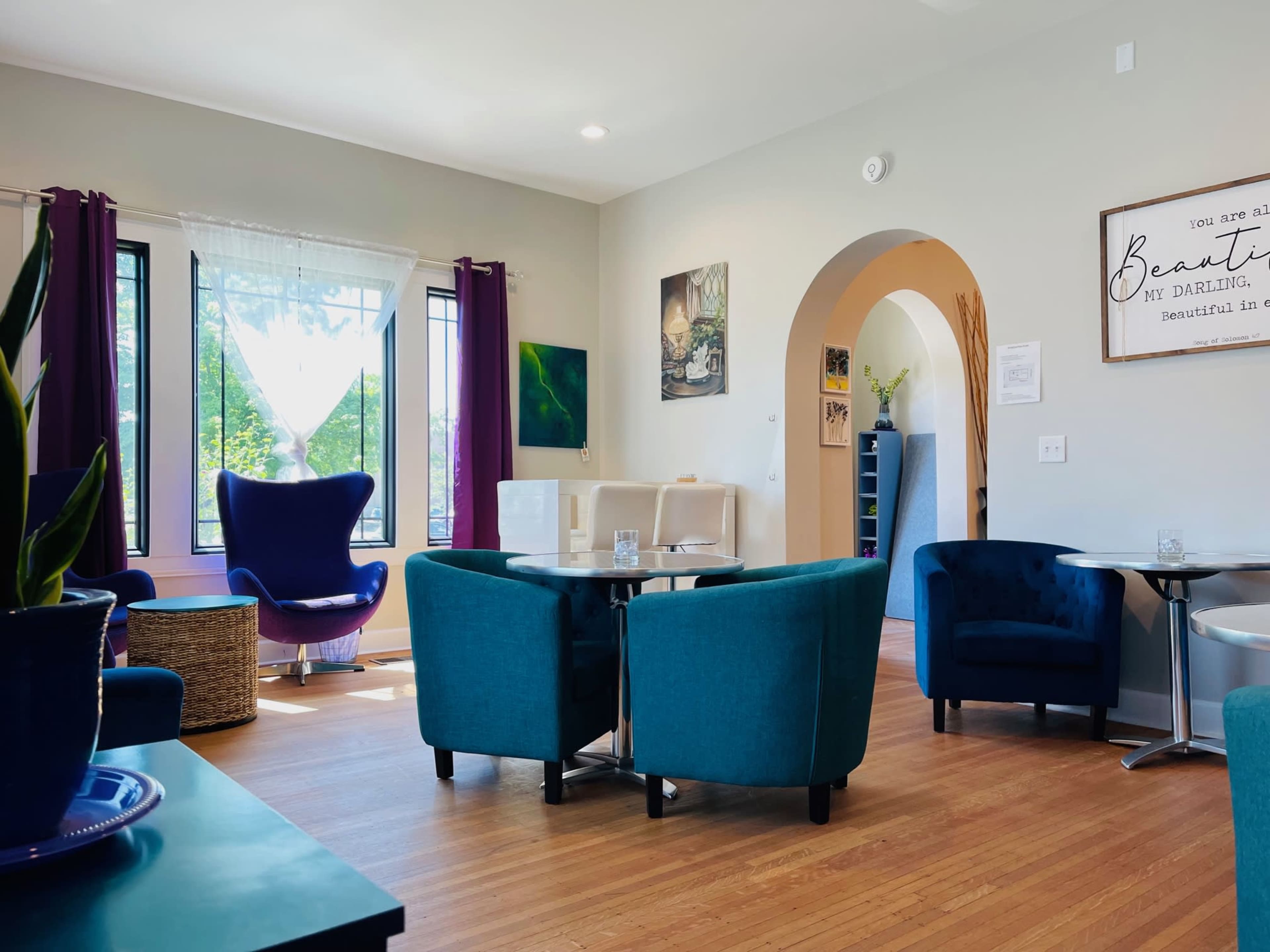 A modern, cozy lounge area with teal chairs and a large purple armchair, featuring large windows and light-colored walls.