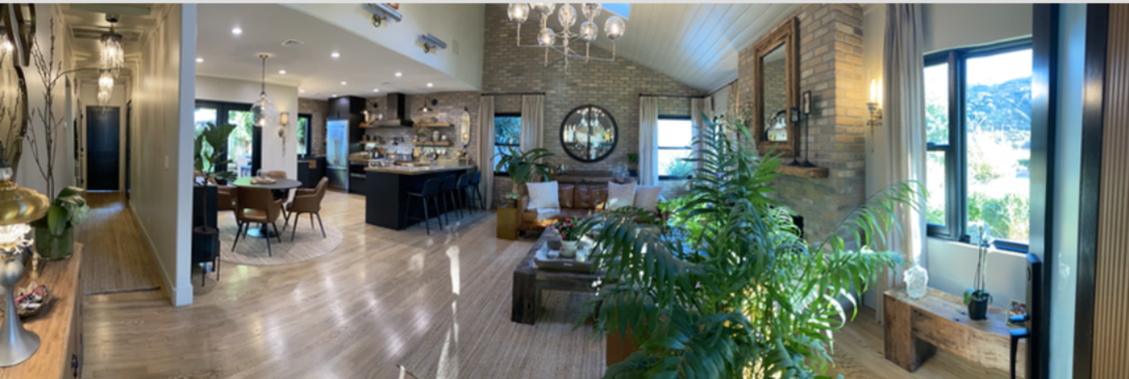 A spacious, modern living area with a kitchen, dining space, and large windows, featuring plants and a mix of natural and artificial lighting.