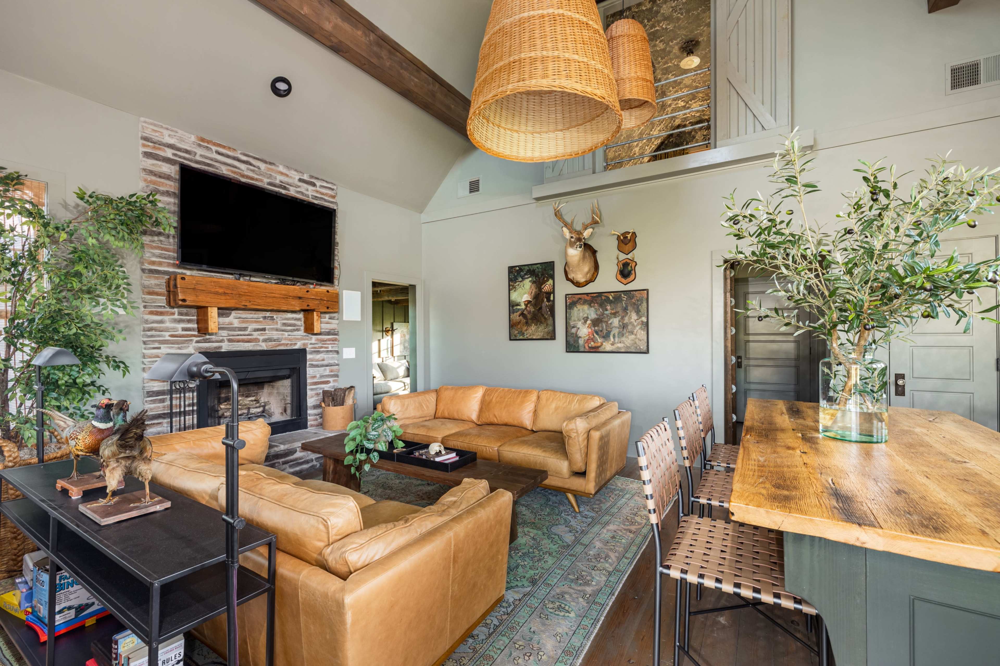The living room features a leather sectional sofa, a stone fireplace, and a wooden dining table with metal chairs, surrounded by greenery and decorative artwork.