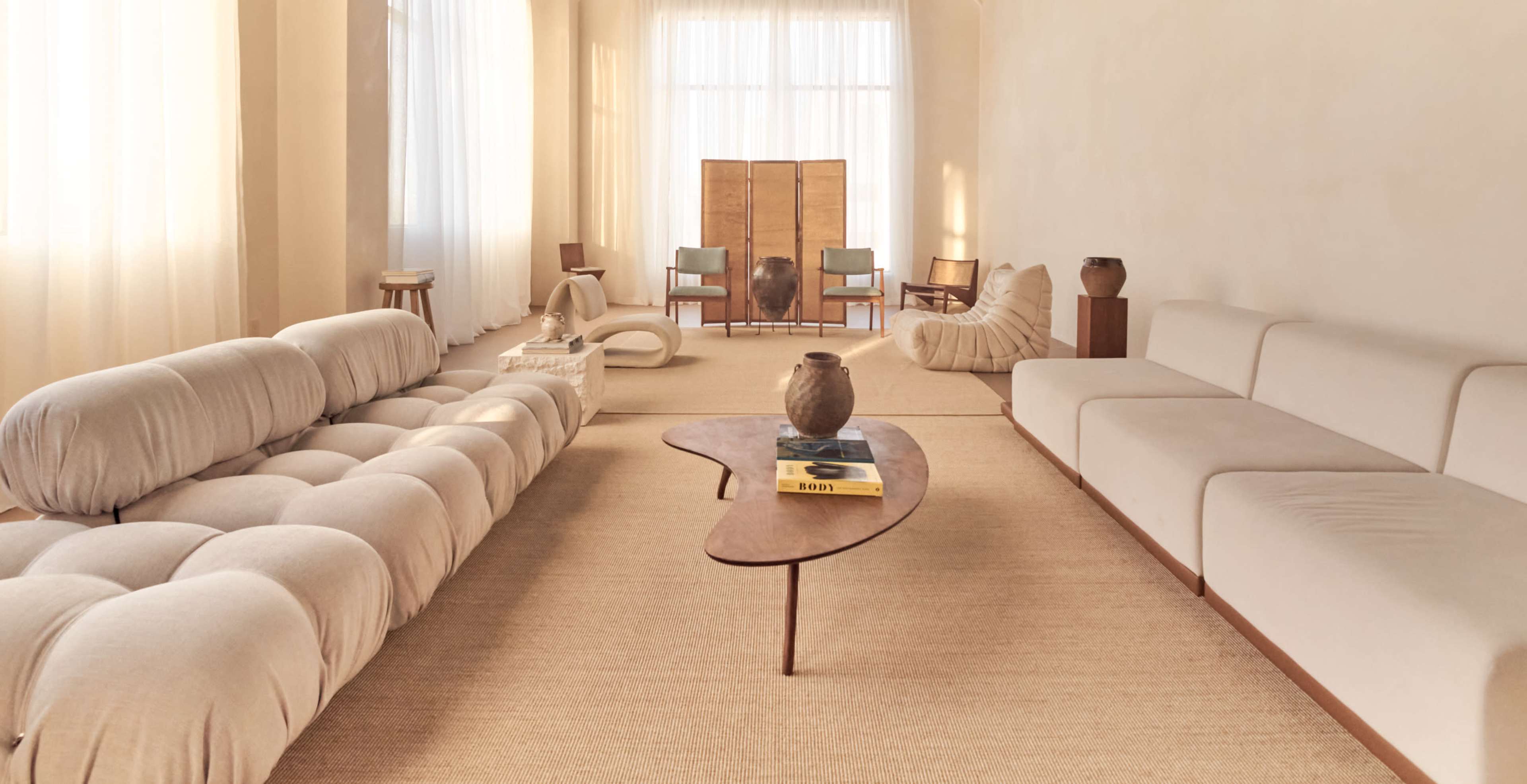 The image shows a minimalist living room with a light-colored interior, featuring a curved sofa, a wooden coffee table, and several pieces of furniture arranged for a spacious feel.