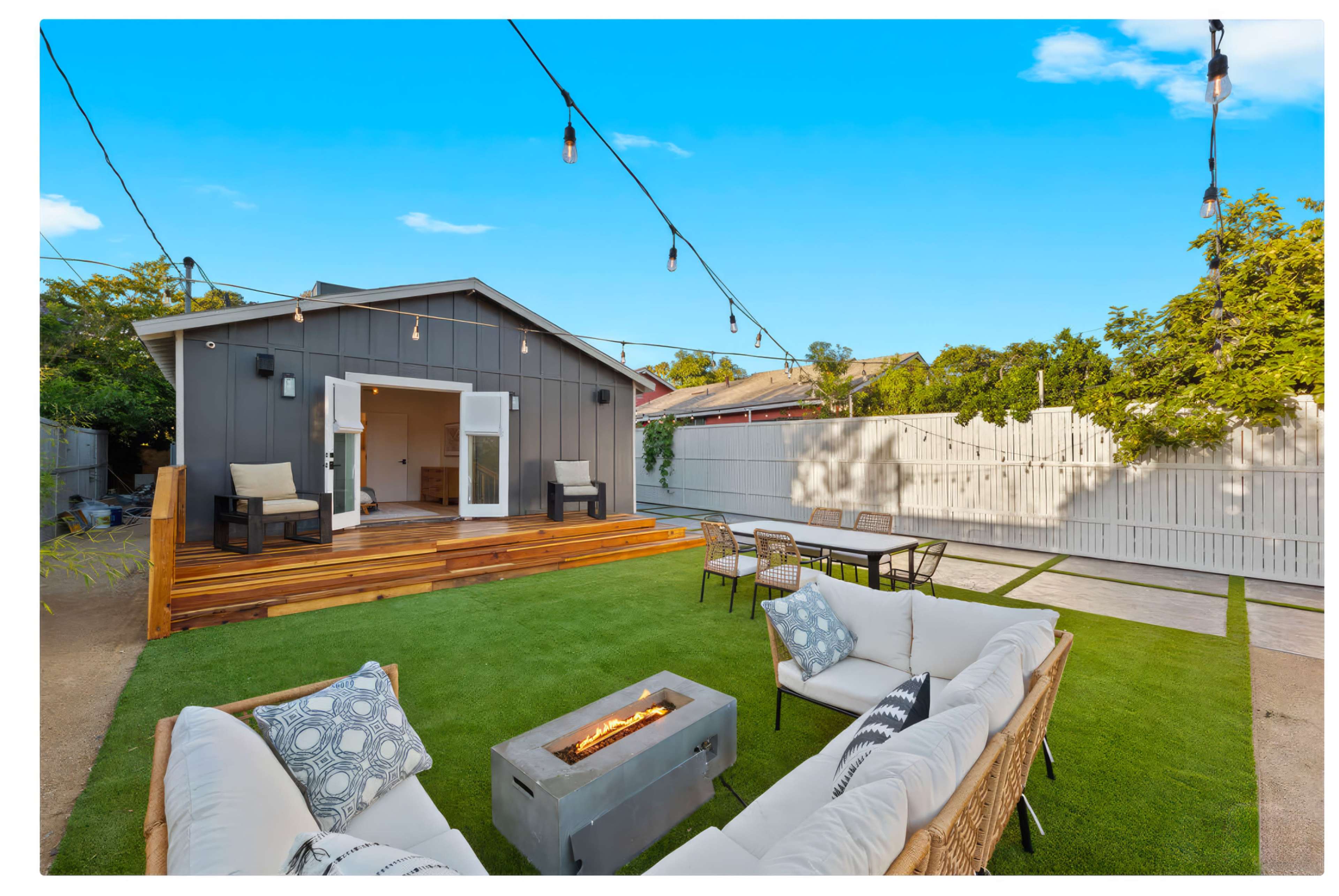 A backyard features a modern gray house with a wooden deck, outdoor seating, a fire pit, and string lights overhead.