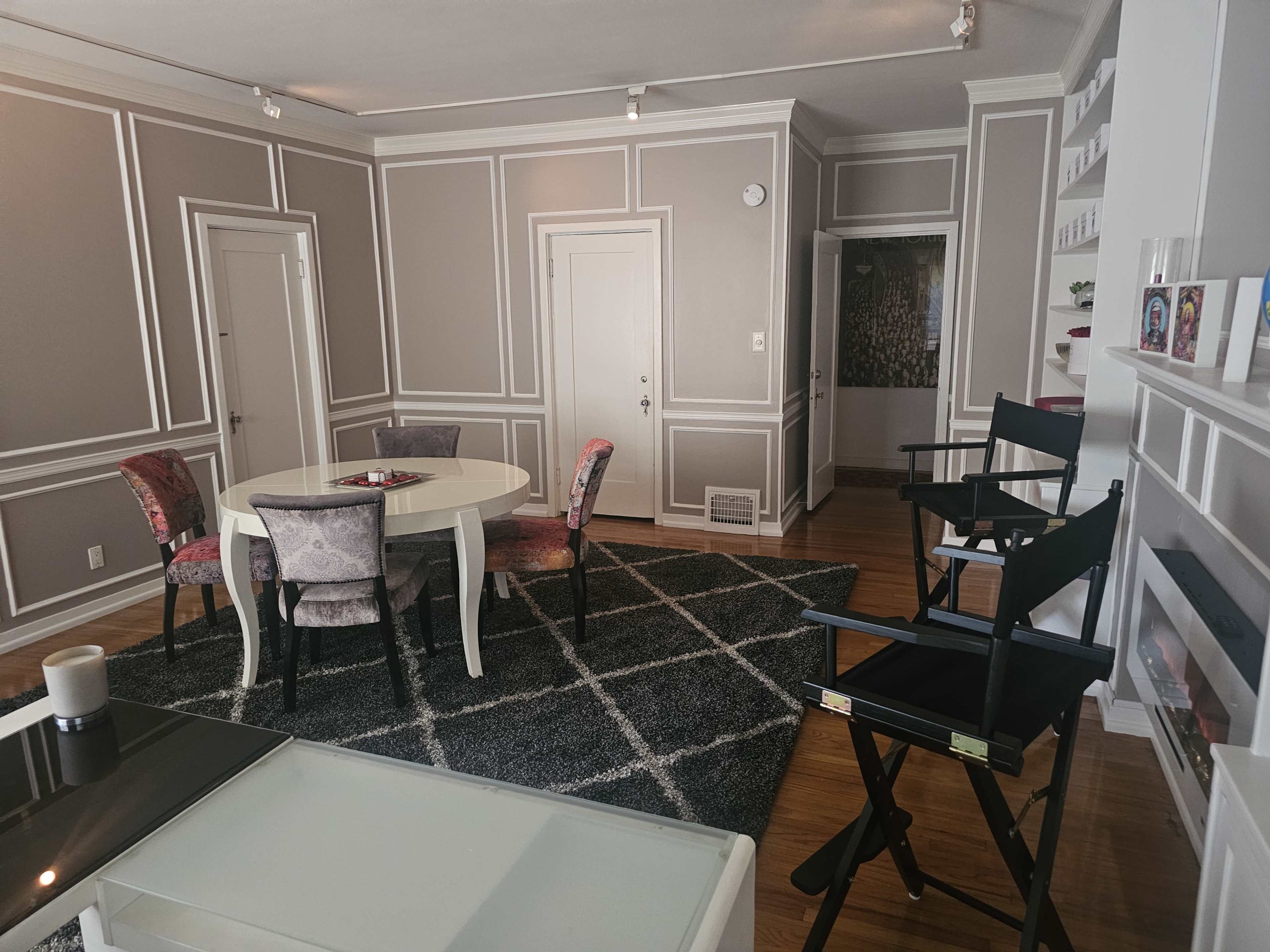 The image shows a modern, well-decorated room featuring a round dining table with chairs, a plush area rug, and a stylish coffee table, along with a small entryway visible in the background.