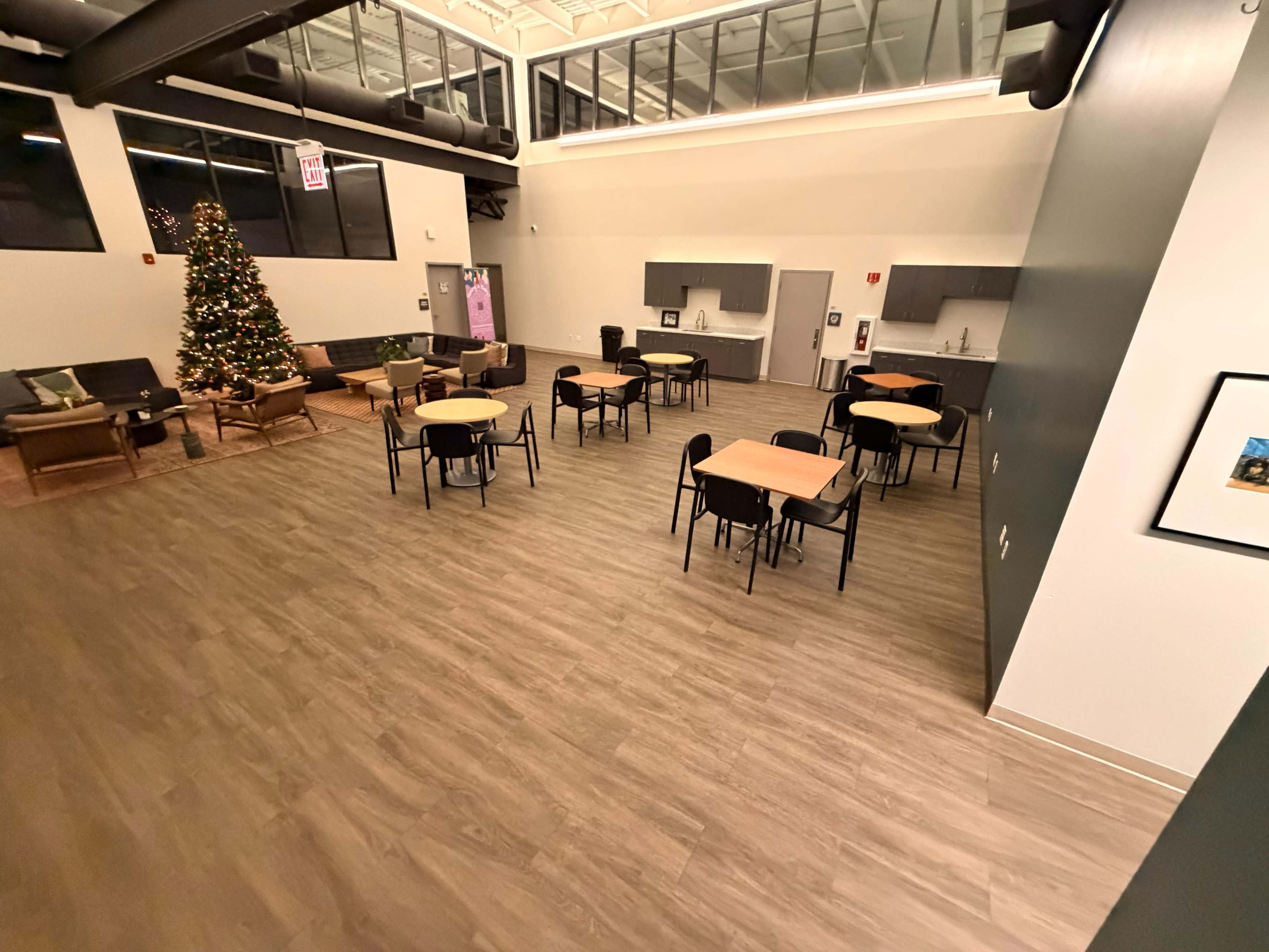 The image shows a spacious indoor area with wooden flooring, several round tables and chairs, a Christmas tree in one corner, and a kitchenette in the background.