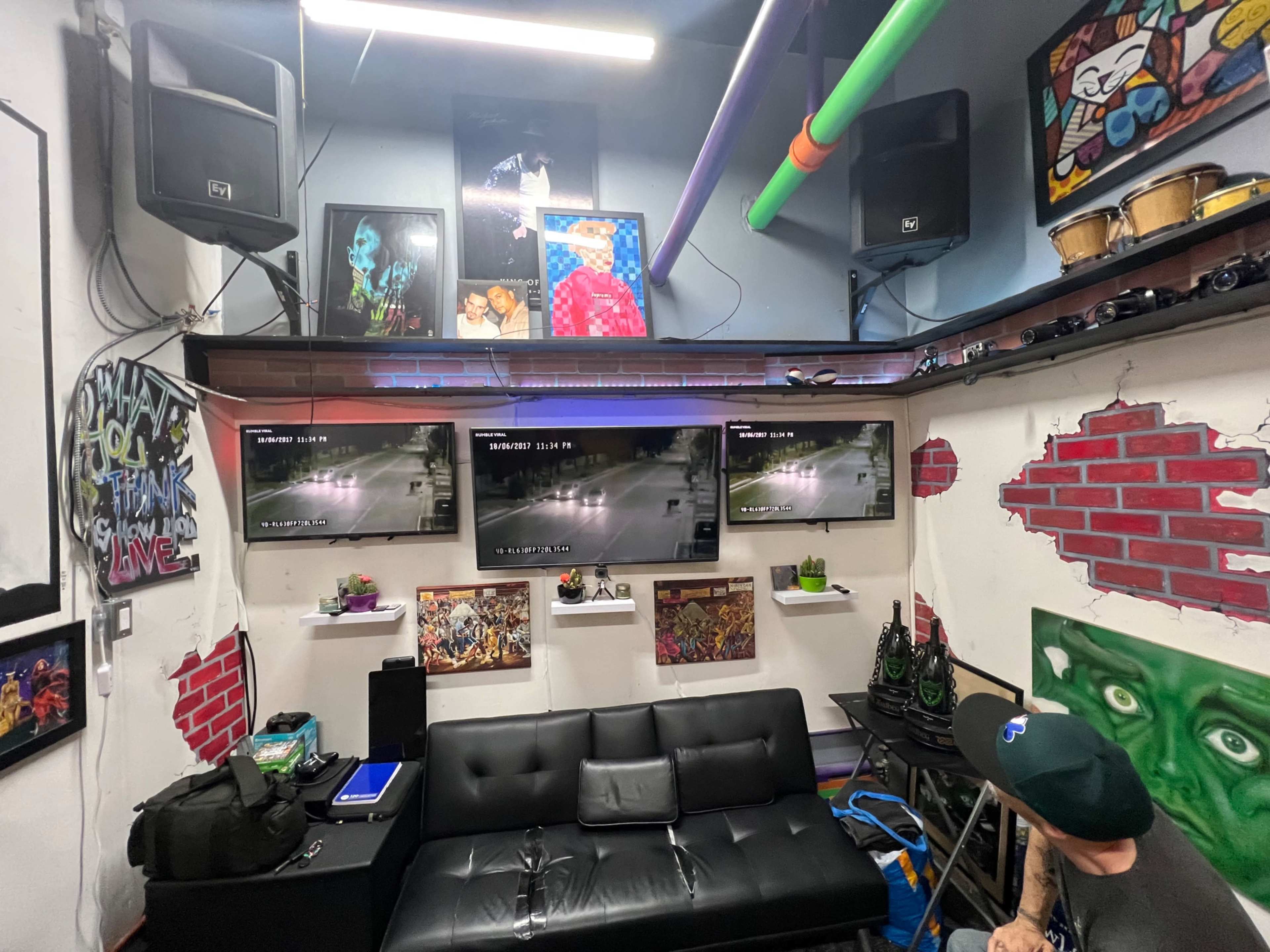 A room with a black sofa, multiple wall-mounted monitors displaying footage, vibrant artwork, and audio equipment.