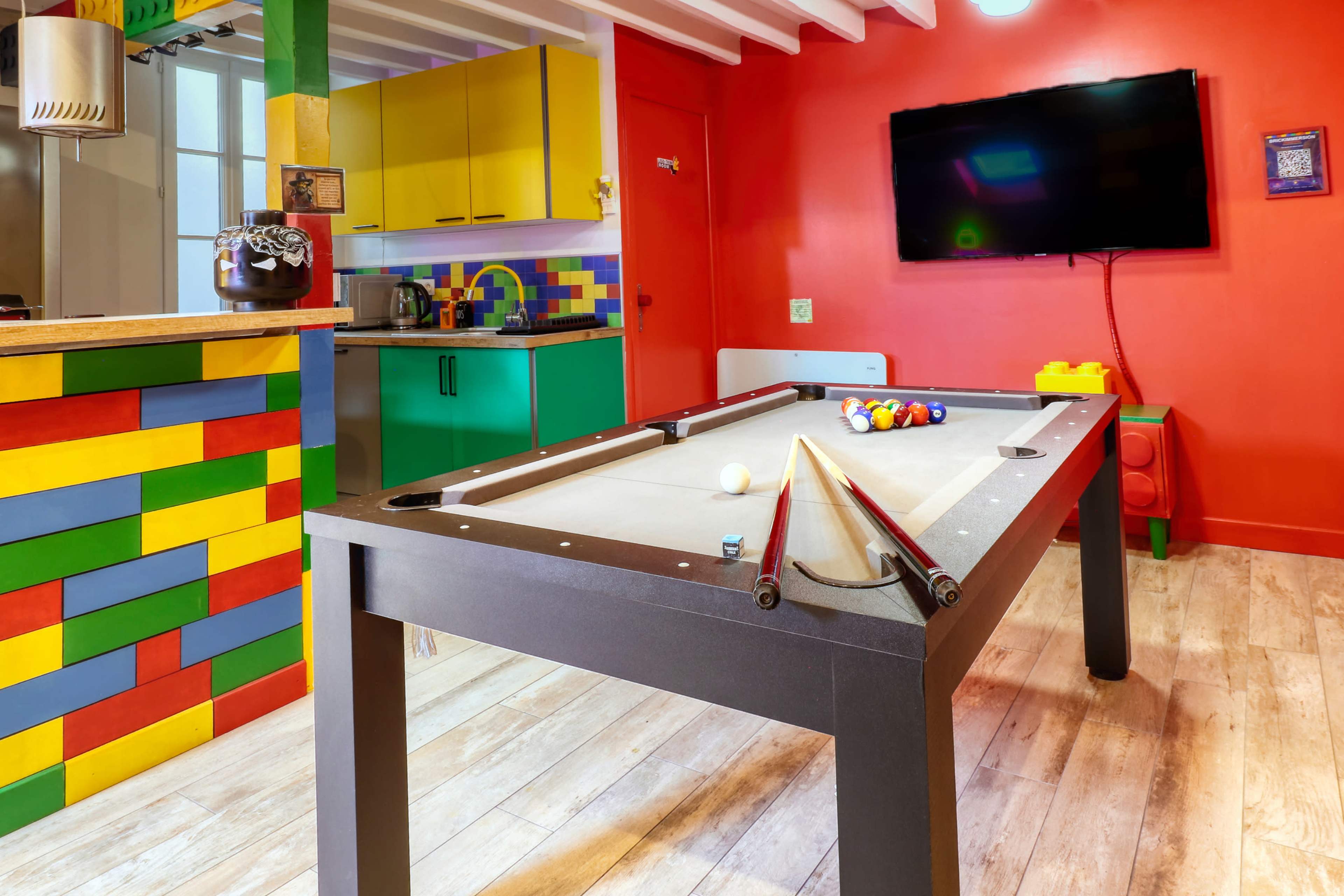 The image shows a colorful game room featuring a pool table, with a vibrant kitchen area in the background and a television mounted on the red wall.