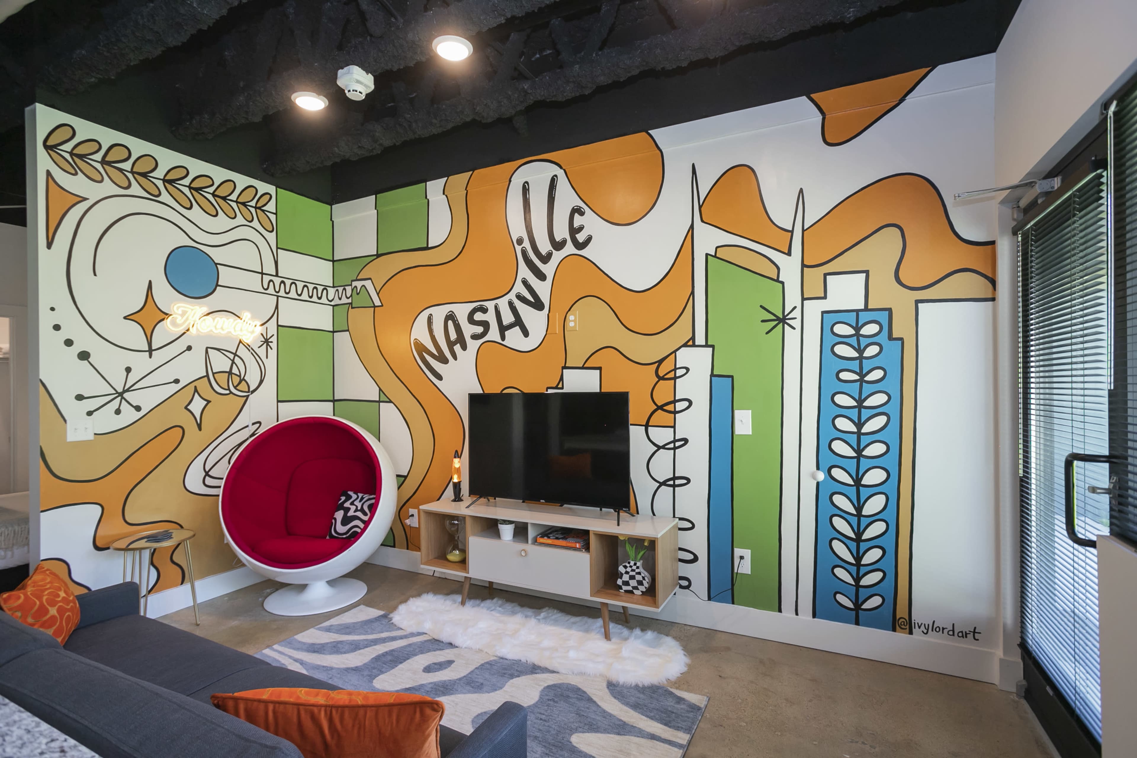 The image shows a modern living area featuring a brightly colored mural on one wall, a red egg-shaped chair, a television on a wooden stand, and a concrete floor.