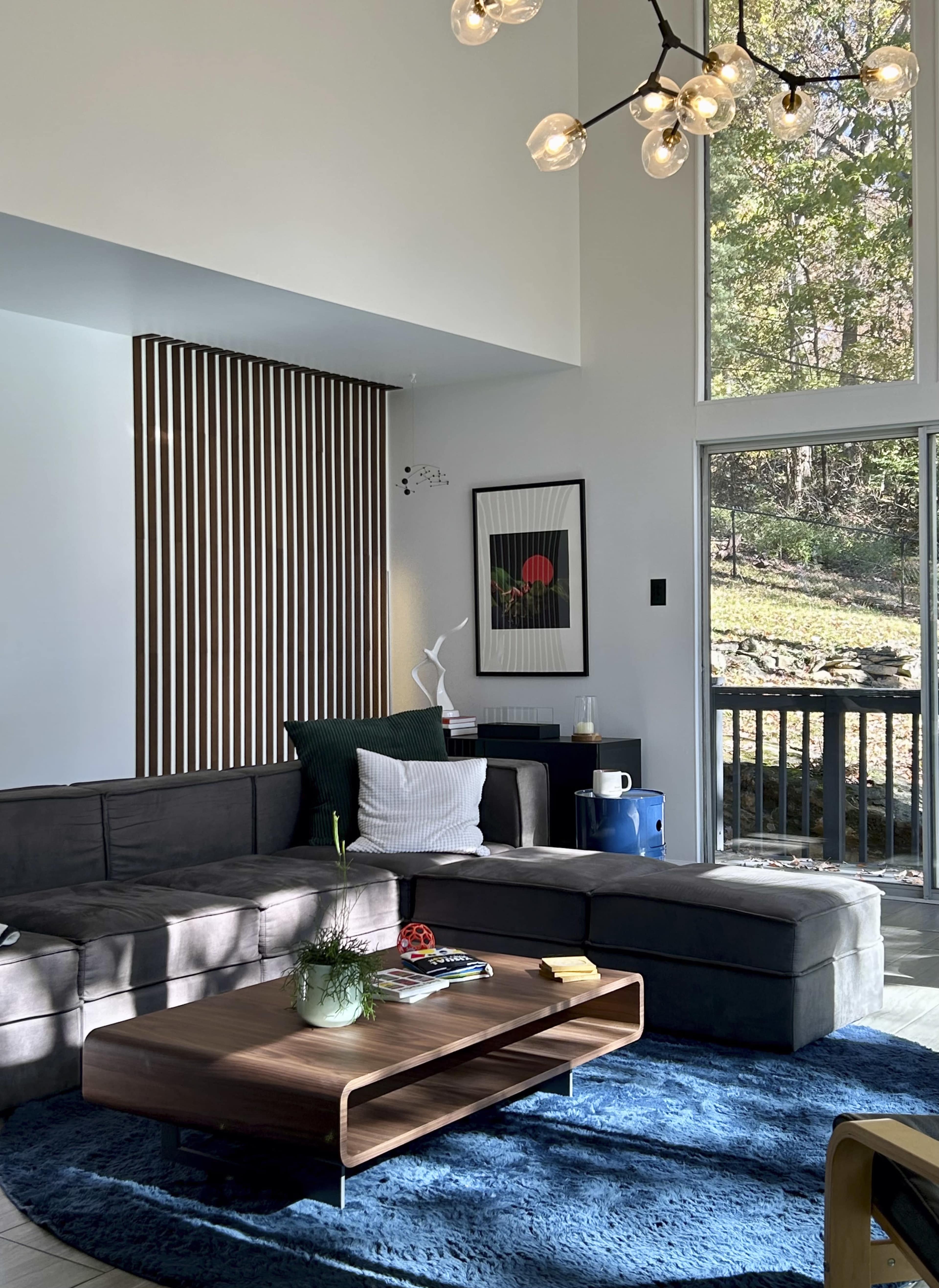 The living room features a sectional sofa, a wooden coffee table on a blue rug, and a large window with a view of trees outside.